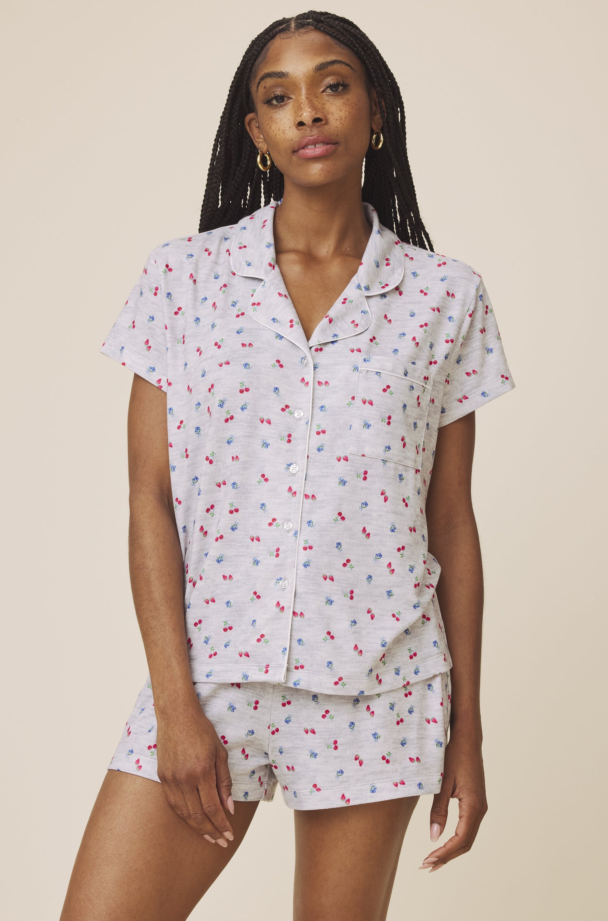 A woman models the Splendid Flora Pillow Soft PJ Short Set in White Multi, featuring a cherry print, button-up top, and matching shorts for a cute, comfortable look against a plain beige background.