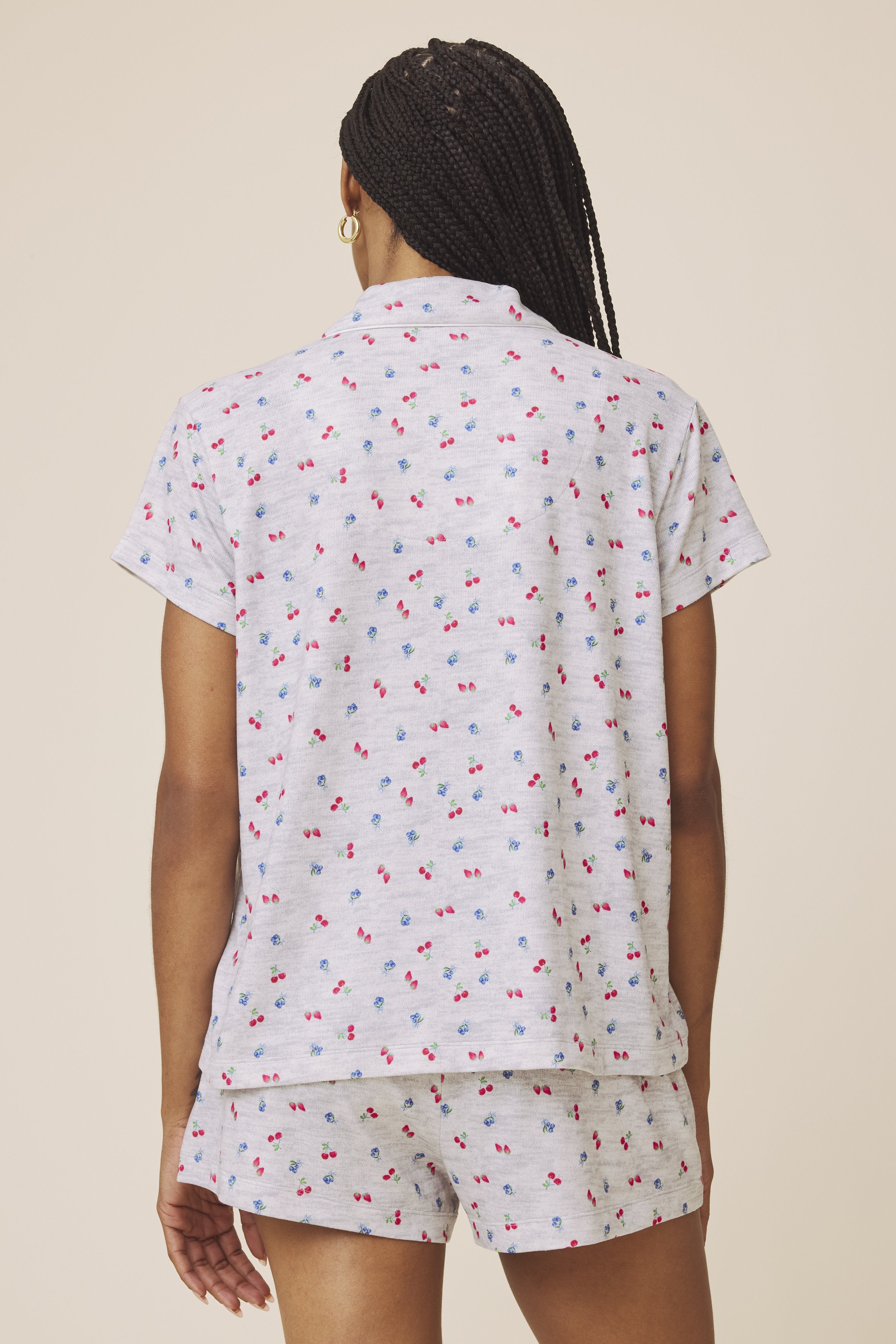 A person with braided hair stands facing away, wearing the Splendid Flora Pillow Soft PJ Short Set: a light gray short-sleeved top and matching shorts featuring a small red and blue cherry print.