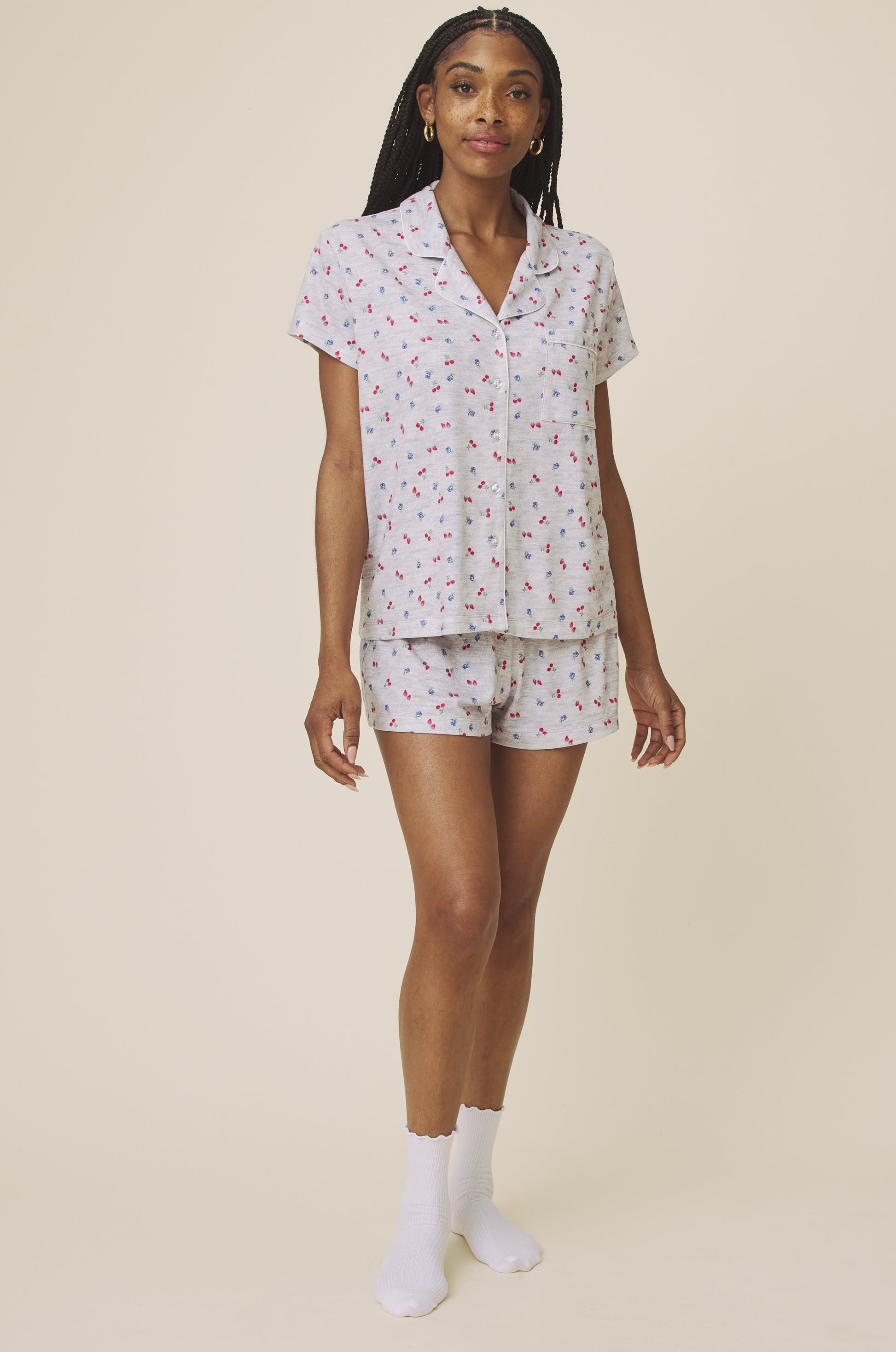 A woman with long braids and a relaxed expression models the Splendid Flora Pillow Soft PJ Short Set, featuring a lightweight light gray pajama set with a colorful print, short sleeves, matching shorts, and white socks against a neutral background.