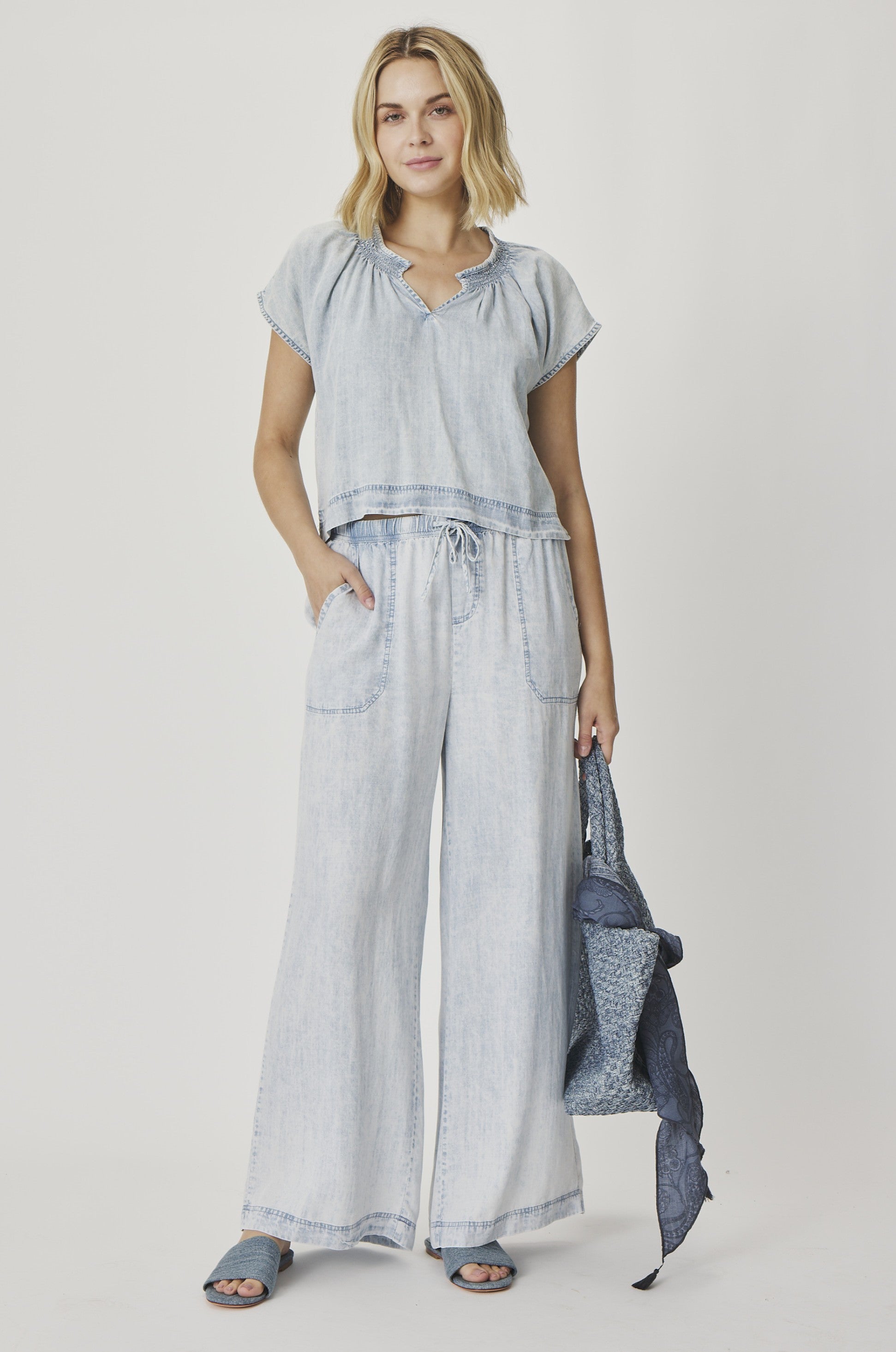 A woman stands against a white background wearing the Splendid Meadow Indigo Blouse, a light blue loose-fitting short-sleeve top with a ruched neckline, paired with matching wide-leg pants, blue sandals, and holding a patterned bag and scarf.
