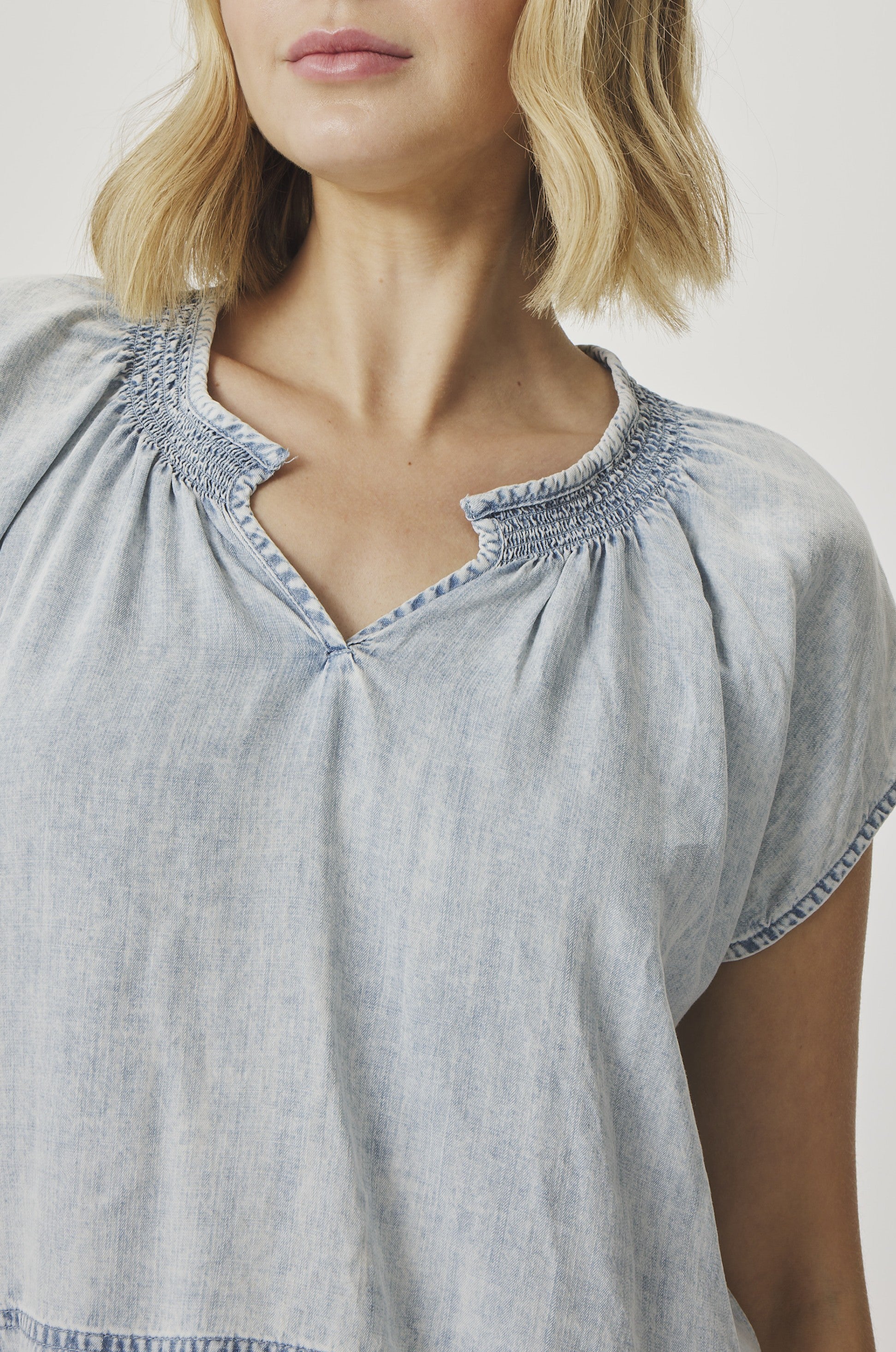 A woman with blonde hair models the Splendid Meadow Indigo Blouse, a light blue, short-sleeved soft denim top with a notched neckline. The photo is cropped from her shoulders to just above her waist.