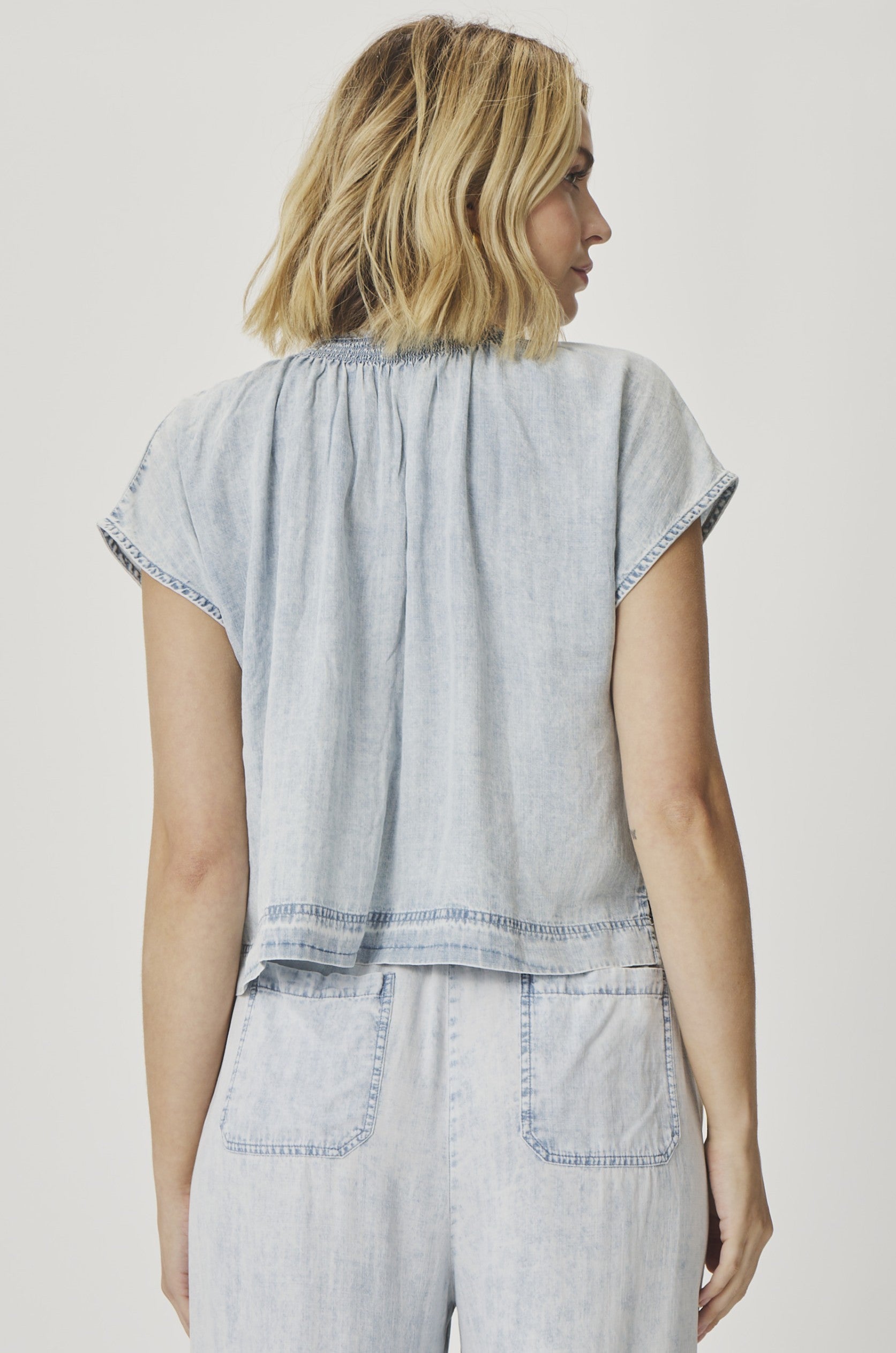 A woman with shoulder-length blonde hair is seen from the back wearing the Splendid Meadow Indigo Blouse—a light blue, soft denim short-sleeve top with a ruched neckline. She pairs it with matching high-waisted cropped denim pants against a plain white background.