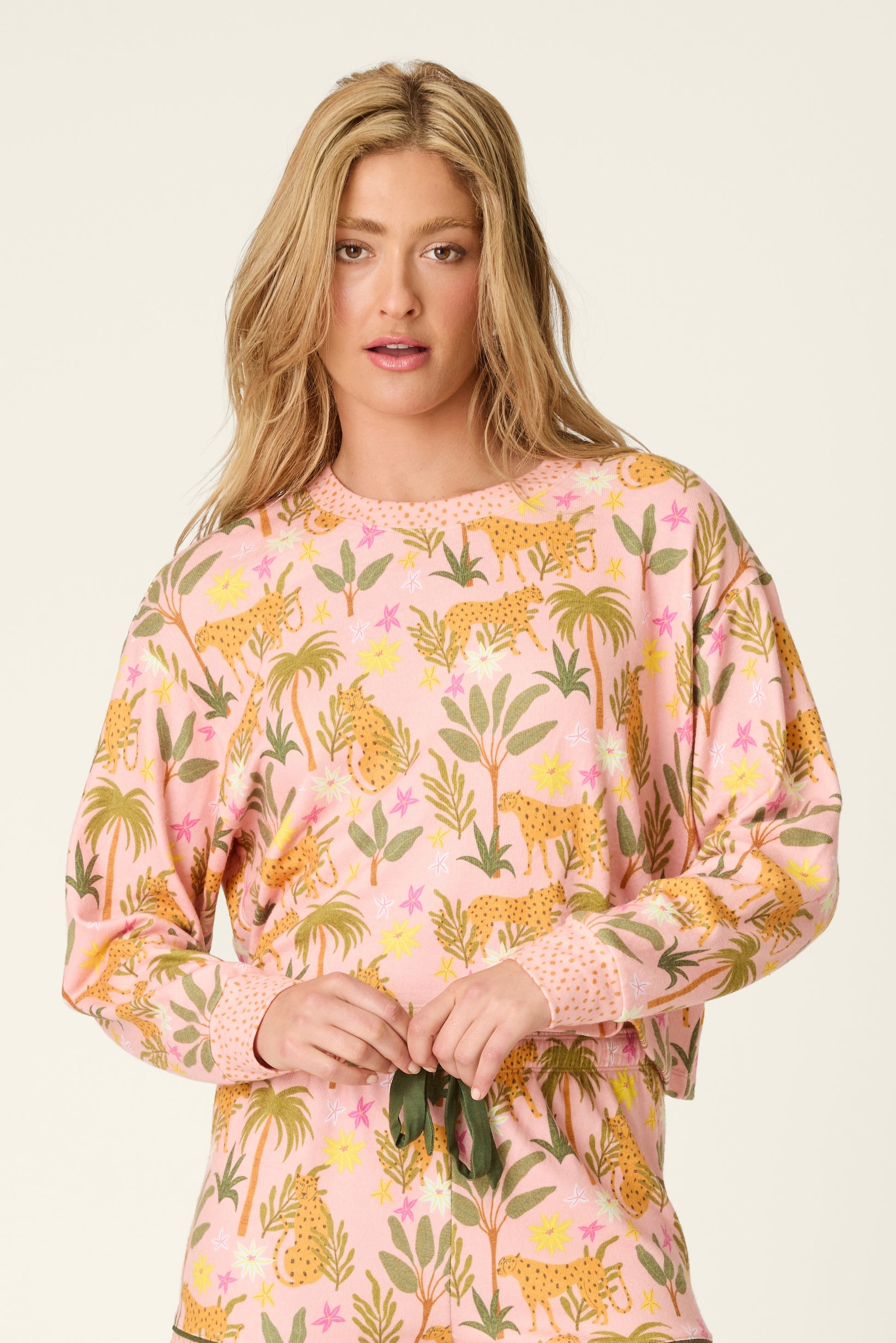 A woman with long blonde hair wears the PJ Salvage In The Jungle Top, a peachy knit pullover featuring a colorful leopard and floral jungle print. She stands against a plain background and looks toward the camera with a neutral expression.