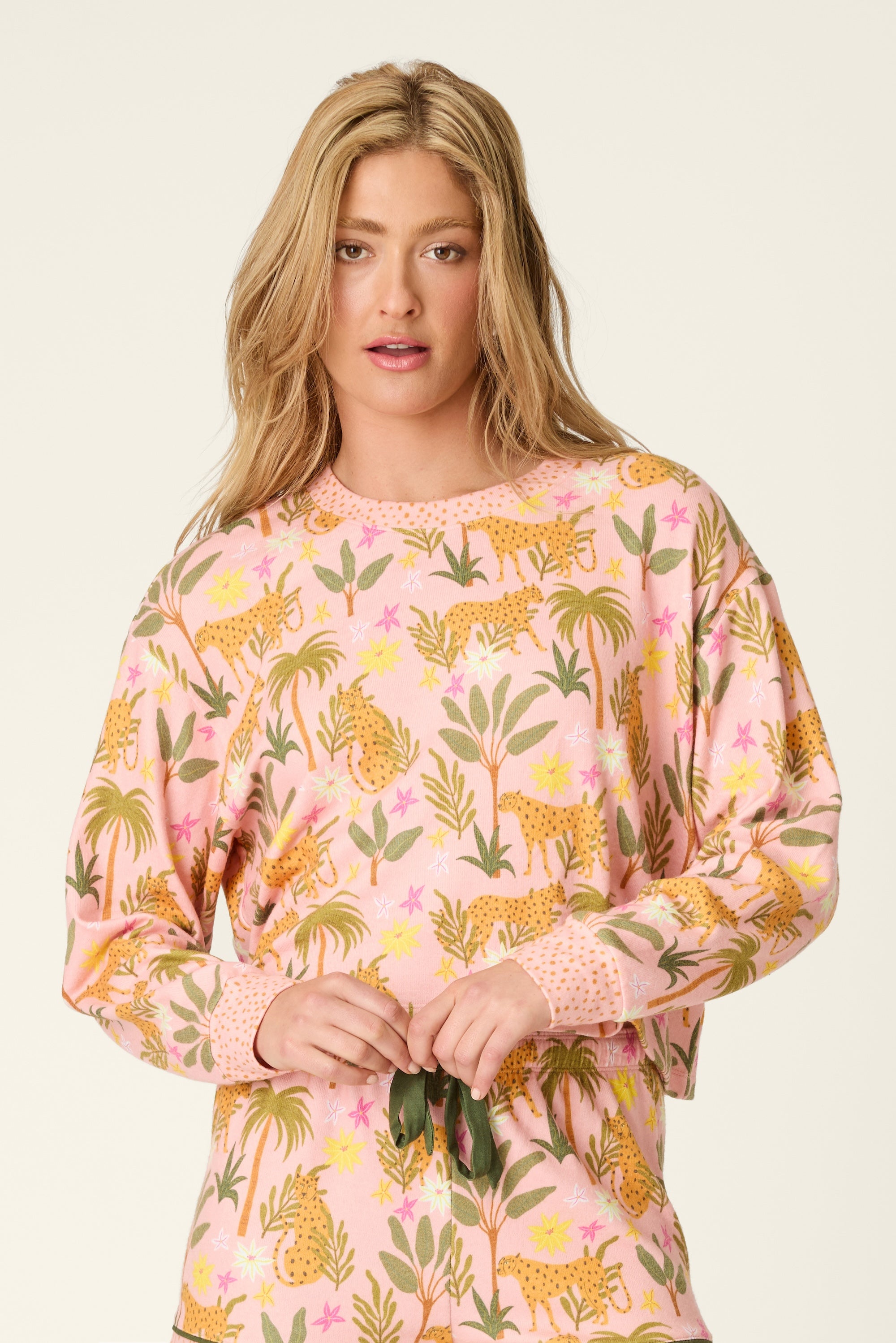 A woman with long blonde hair wears the PJ Salvage In The Jungle Top, a peachy knit pullover featuring a colorful leopard and floral jungle print. She stands against a plain background and looks toward the camera with a neutral expression.