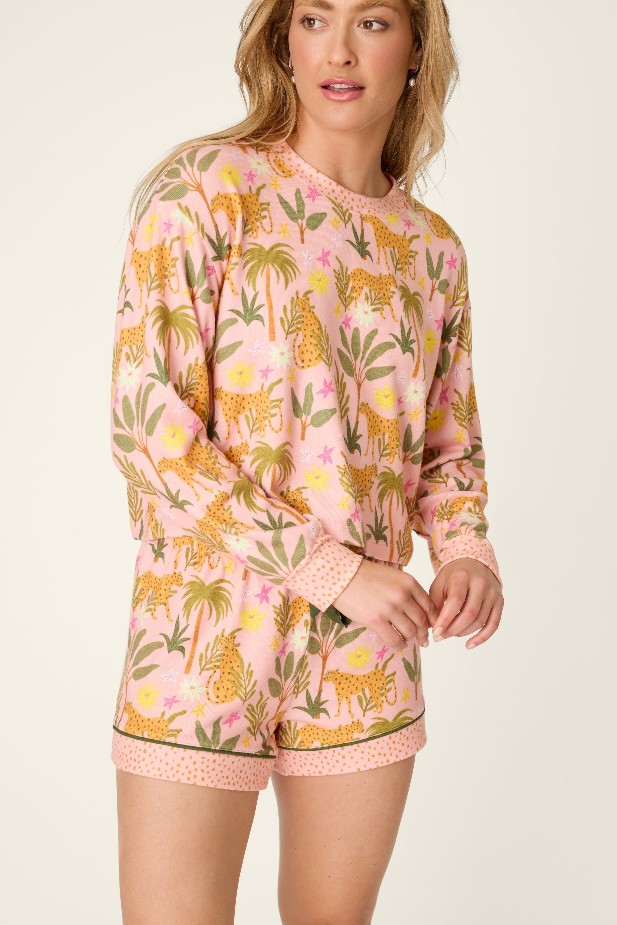 A woman stands looking to the side, wearing the PJ Salvage In The Jungle Top—a long-sleeved pullover pajama with a matching pink jungle print featuring cheetahs and plants—paired perfectly with coordinating shorts finished with a dotted trim.