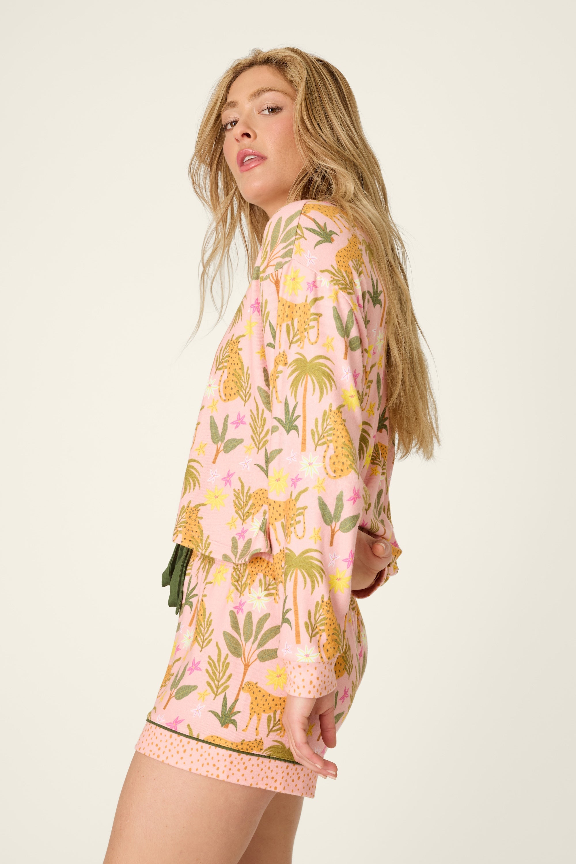 A woman with long blonde hair stands sideways and looks at the camera against a plain background, wearing PJ Salvage’s *In The Jungle Top*—a pink pajama top with a yellow and green jungle print—paired with a cozy peach-toned knit pullover.