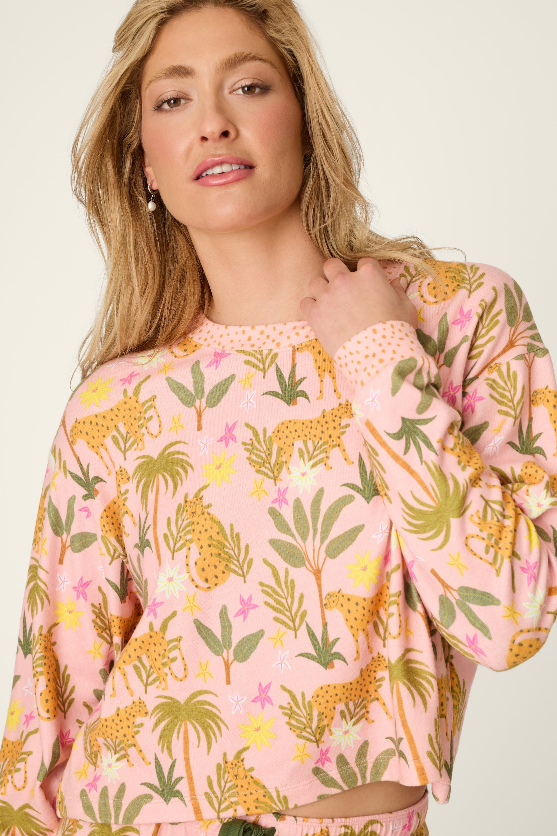 A woman with wavy blonde hair wears the PJ Salvage "In The Jungle Top" in BLOSSOM, a peachy knit pullover featuring a vibrant cheetah and tropical jungle print, looking at the camera with a relaxed expression.