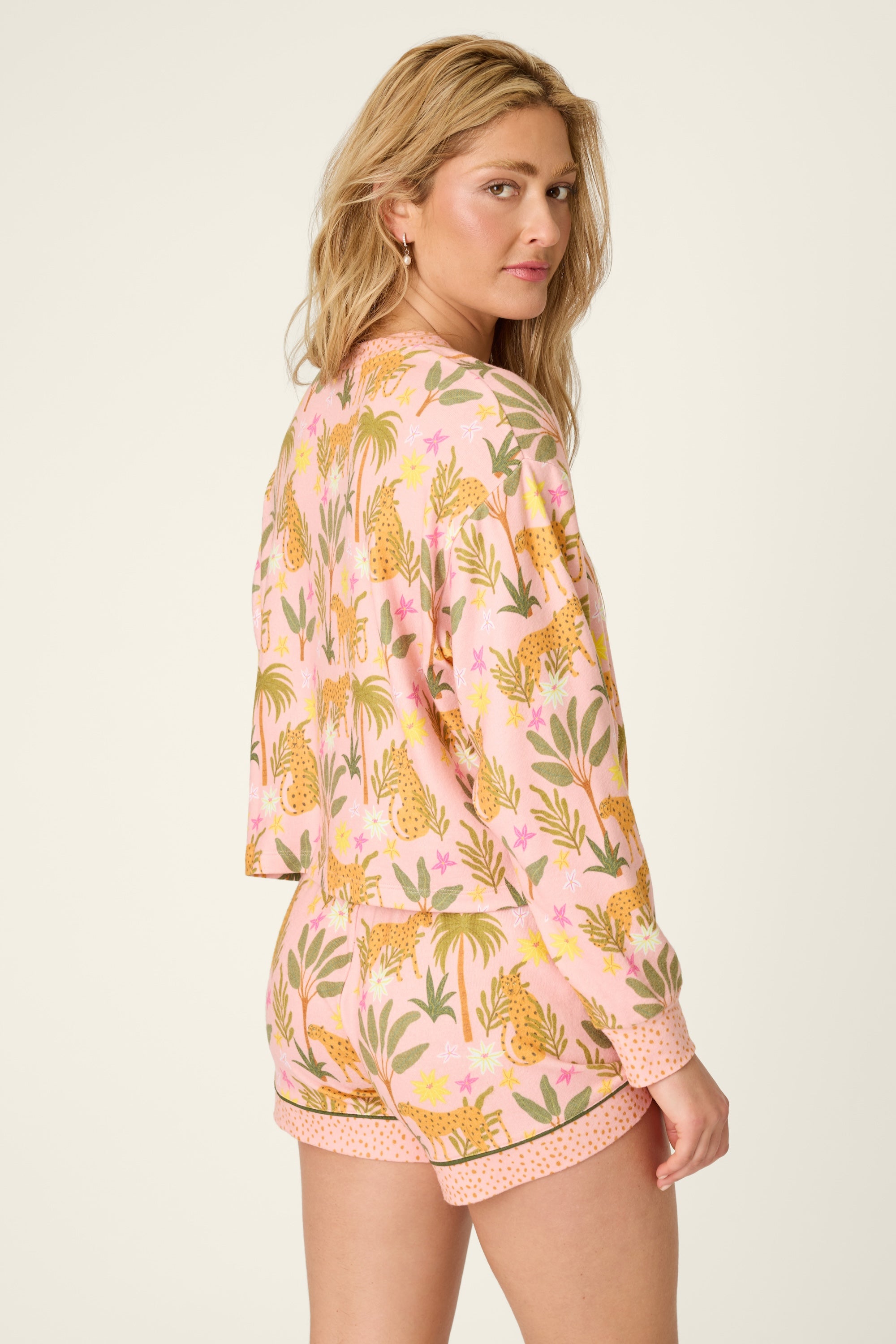 A woman wearing the PJ Salvage "In The Jungle Top," a pink and yellow jungle print long-sleeve pajama top paired with matching shorts, poses looking over her shoulder against a plain light background.