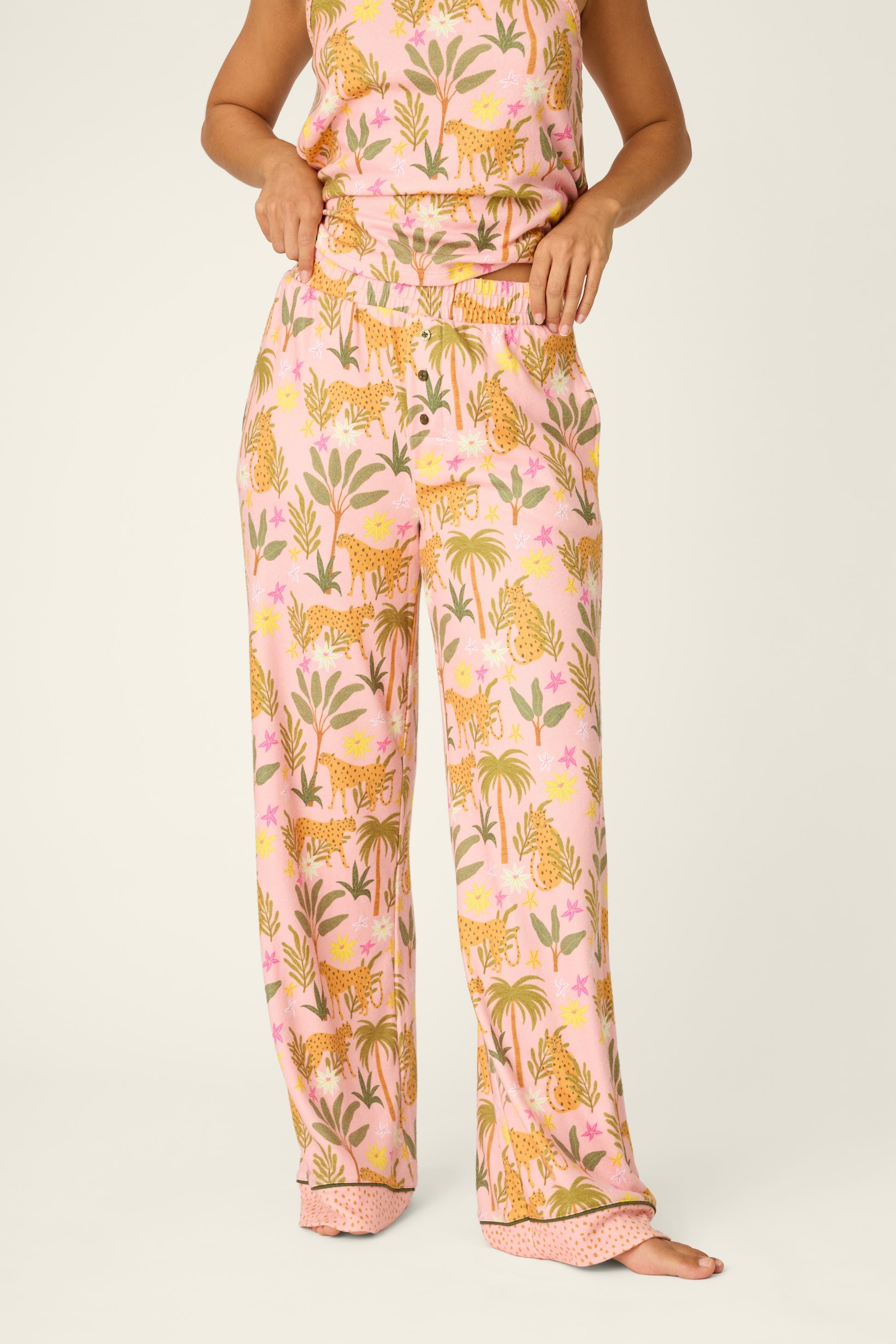 A person stands barefoot against a plain background, hands resting on the elastic waistband of the PJ Salvage In The Jungle Pant in BLOSSOM—a pair of women’s pajama pants featuring a vibrant pattern of tigers and plants.