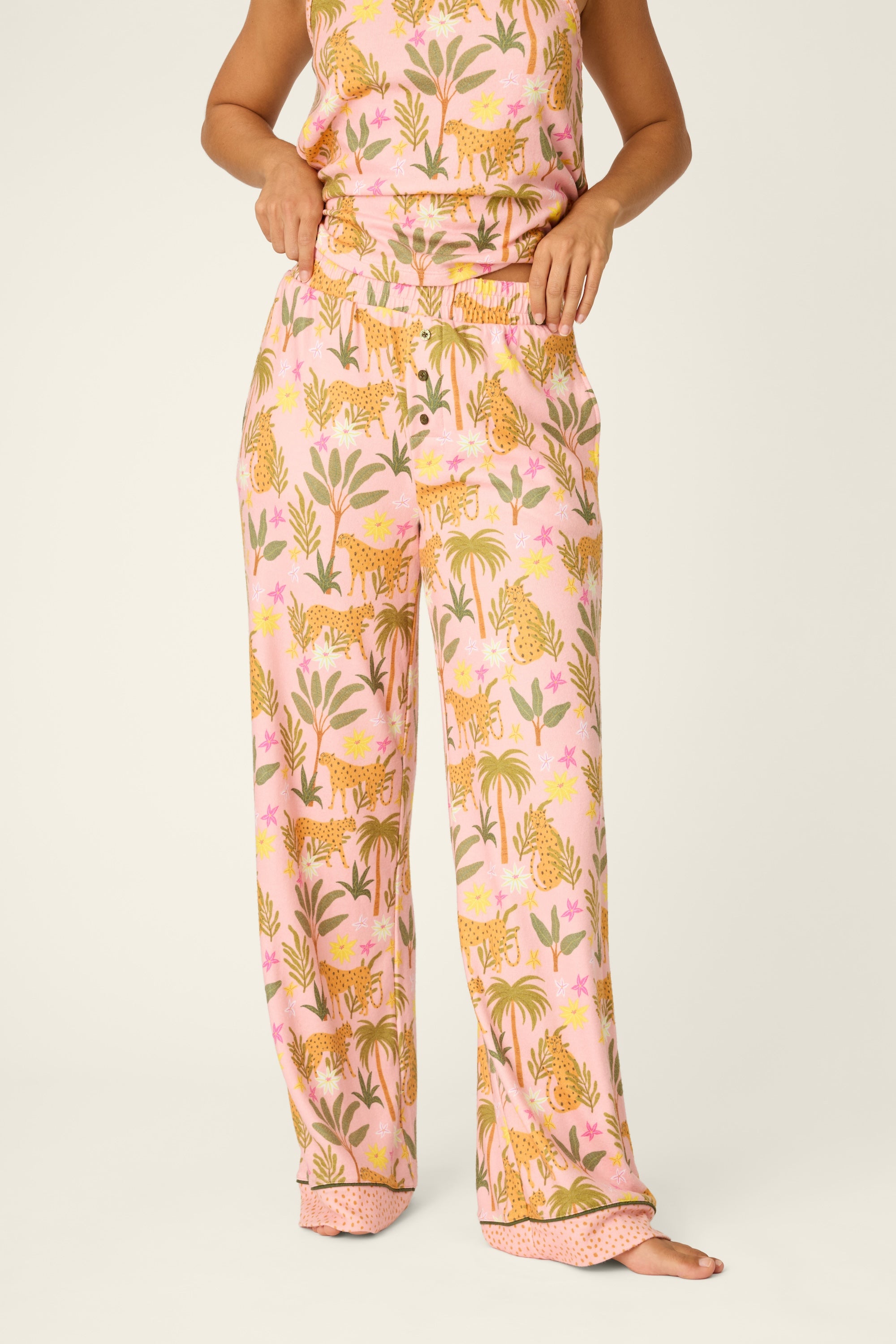 A person stands barefoot against a plain background, hands resting on the elastic waistband of the PJ Salvage In The Jungle Pant in BLOSSOM—a pair of women’s pajama pants featuring a vibrant pattern of tigers and plants.