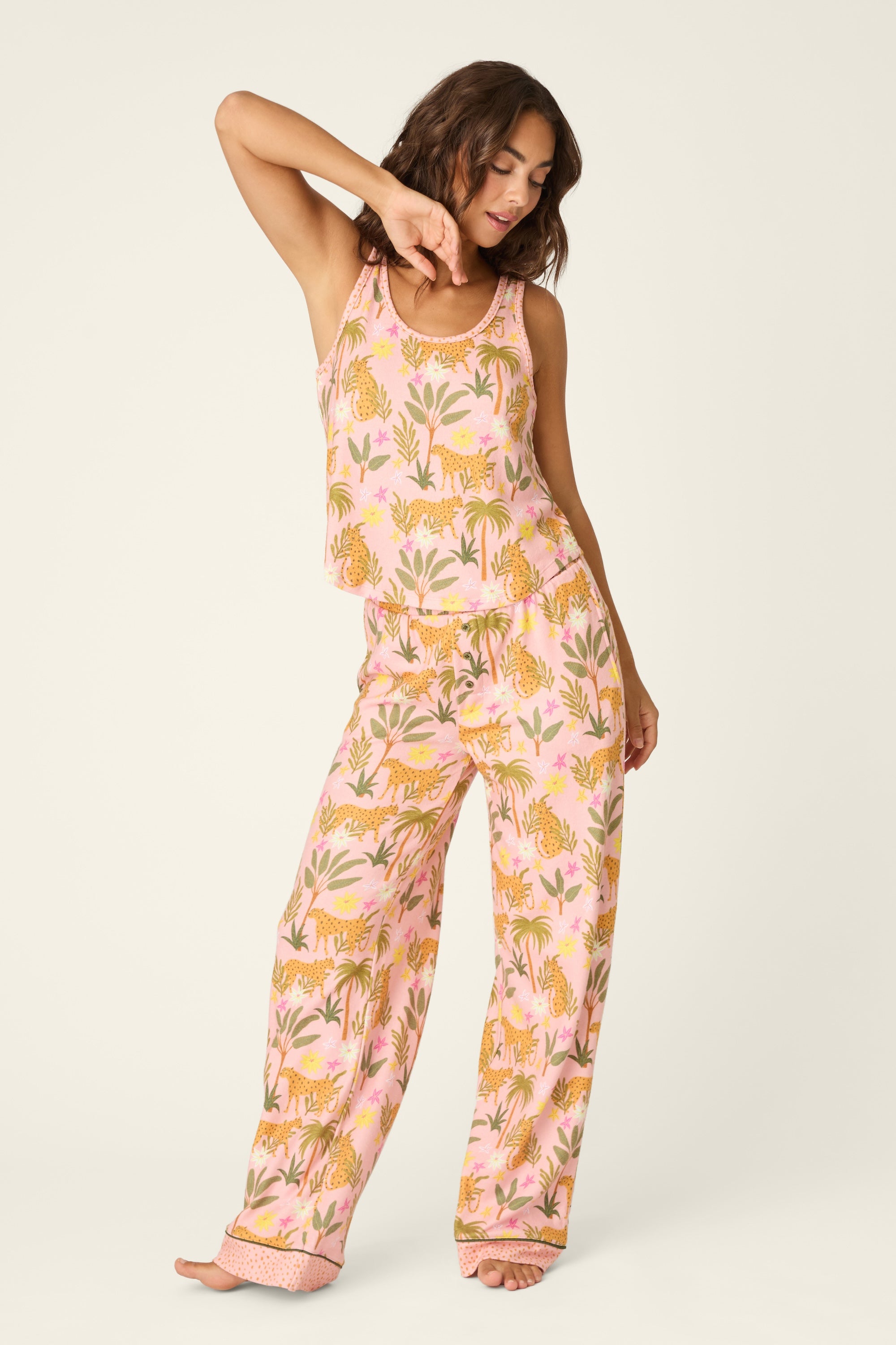 A woman stands barefoot against a plain light background, posing with one arm raised and her head tilted slightly, wearing PJ Salvage’s In The Jungle Pant—a comfortable women’s pajama pant featuring a fun pink design with orange tiger and green leaf prints and an elastic waistband.