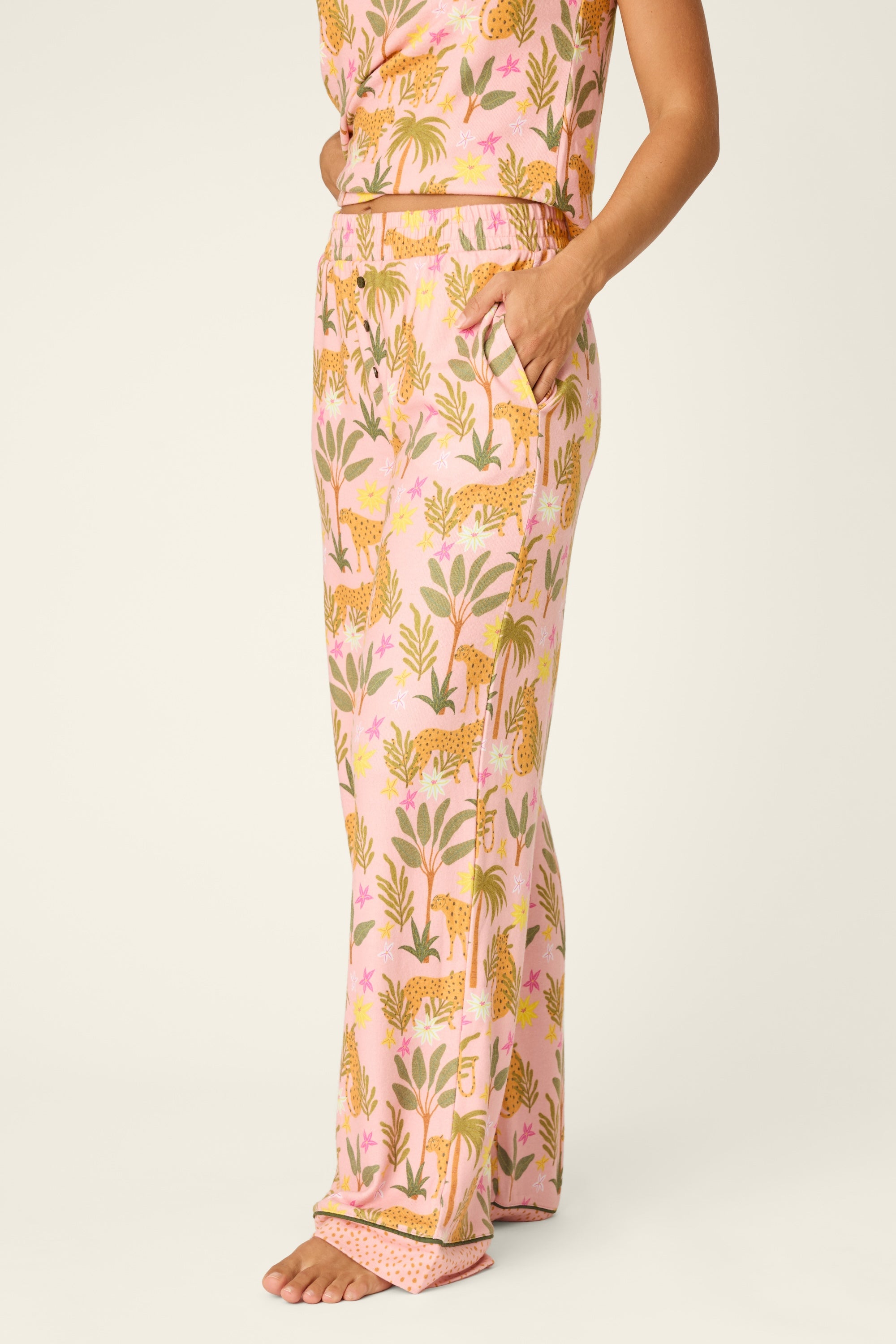 A person stands barefoot against a plain light background, wearing PJ Salvage's In The Jungle Pant—a pair of women's pajama pants with a colorful tropical and leopard print, an elastic waistband, and one hand in their pocket.