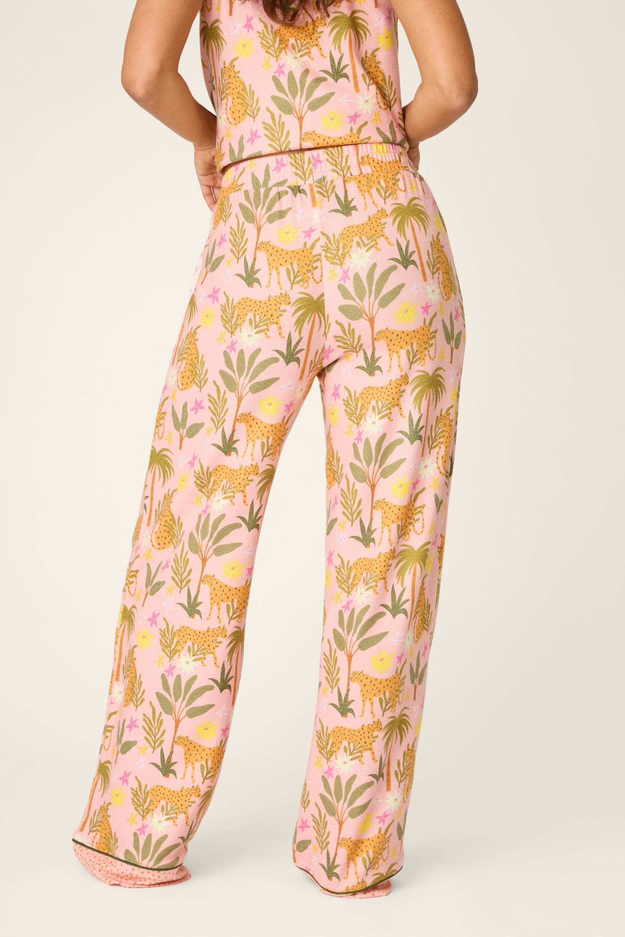 A person seen from the back is wearing PJ Salvage's In The Jungle Pant—women’s pajama pants with an elastic waistband in pink, adorned with playful cheetahs and leafy plants. These loose-fitting pants feature a colorful, jungle-inspired design for a fun and comfortable look.