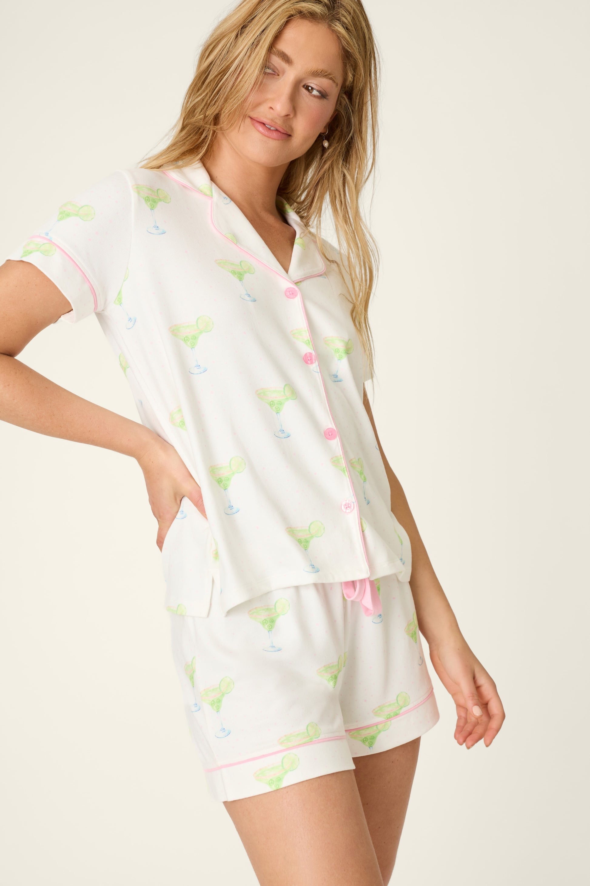 A woman models the PJ Salvage Spicy Marg Pj Set in IVORY, featuring matching women's shorts (2.75" inseam) and a short-sleeve button-up top in soft Mid Butter Jersey with a light margarita glass print and pink trim, against a plain background.