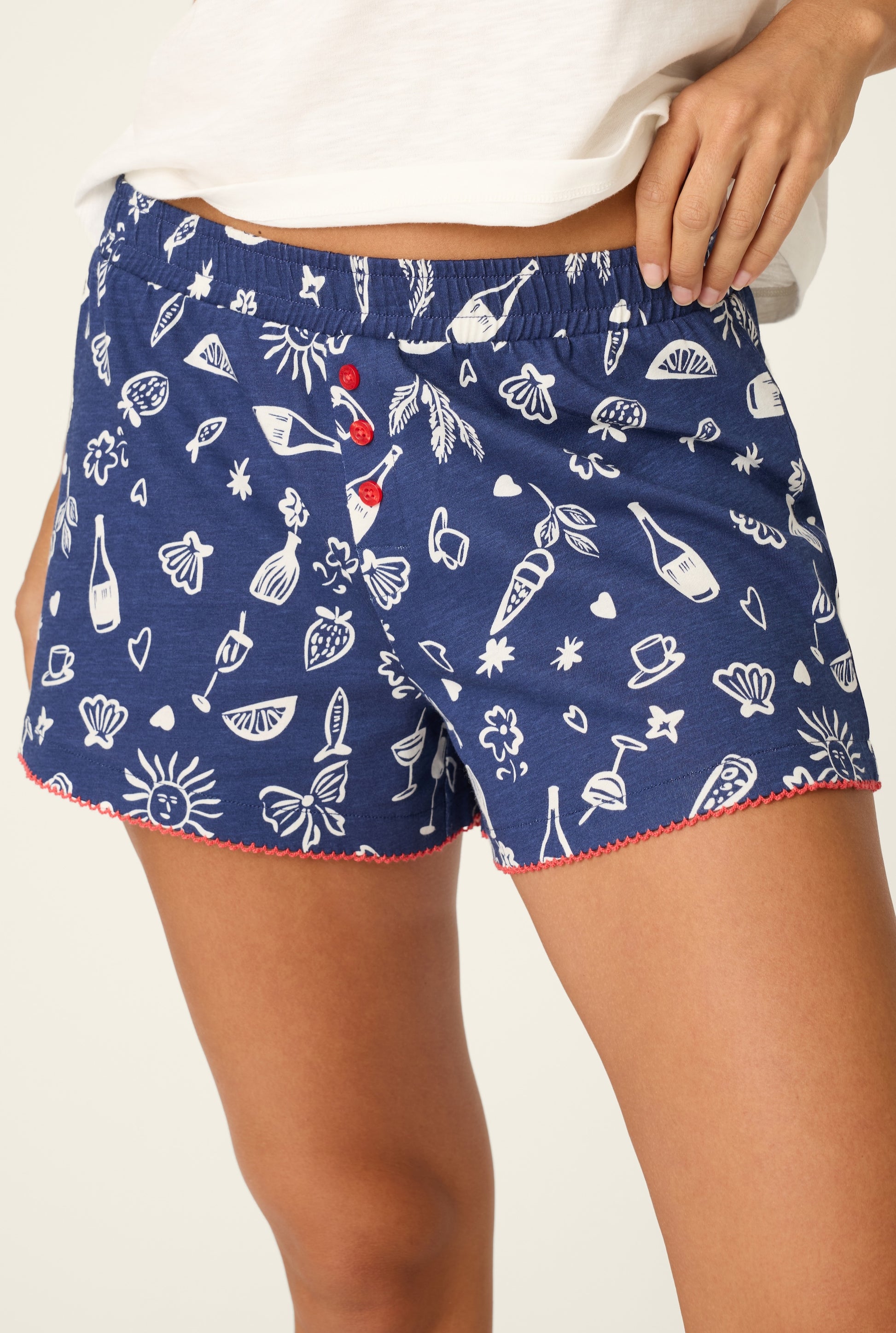 A person wearing PJ Salvage Playful Print Boxer Shorts in navy blue with white fruit, drink, and flower prints, a logo waistband, red buttons, red stitched hem, and part of a white shirt visible.