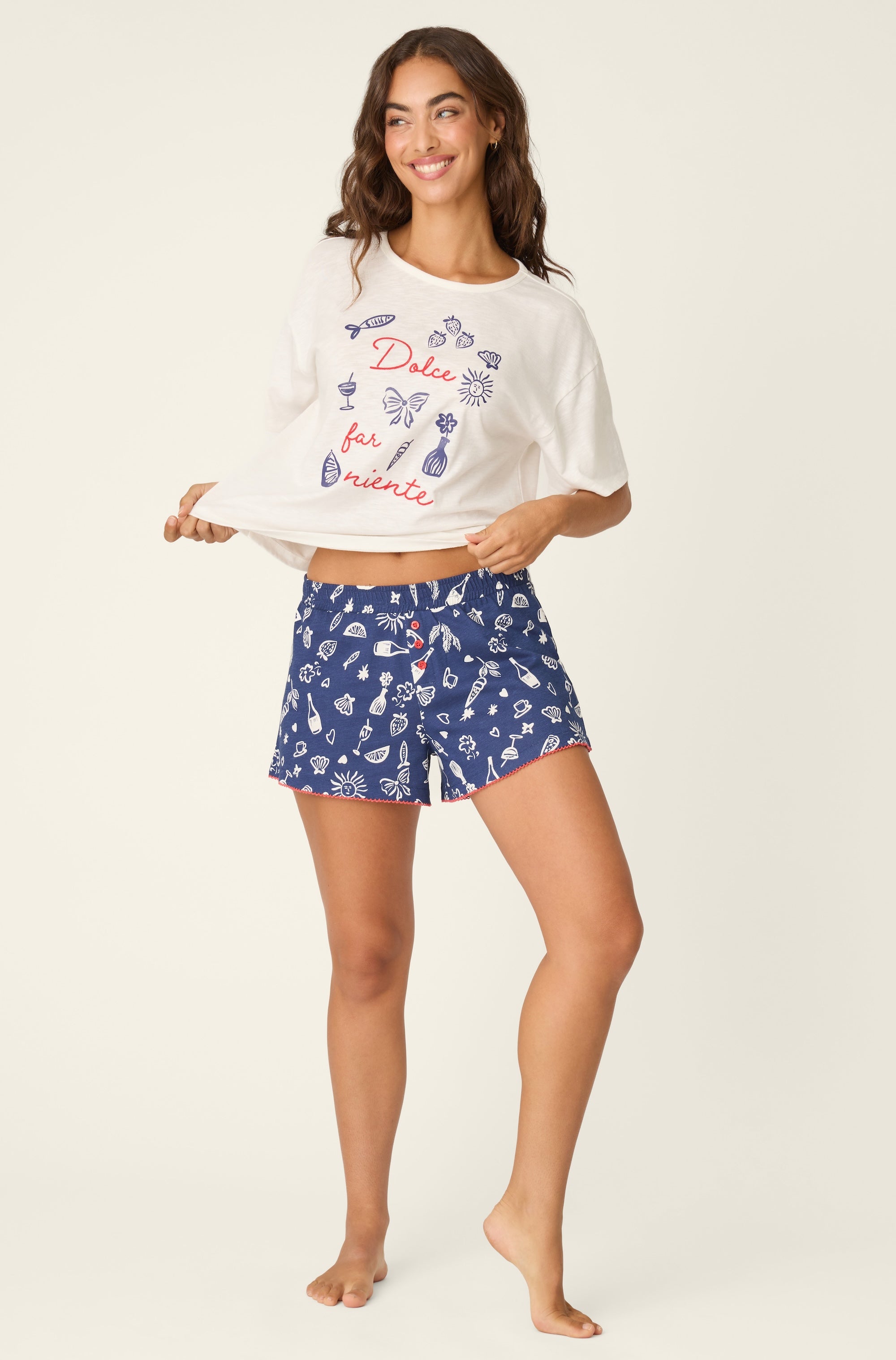 A woman smiles barefoot, holding up her white “Dolce far niente” T-shirt and wearing PJ Salvage Playful Print Boxer Shorts, which feature a logo waistband with blue and white patterns on a light background.