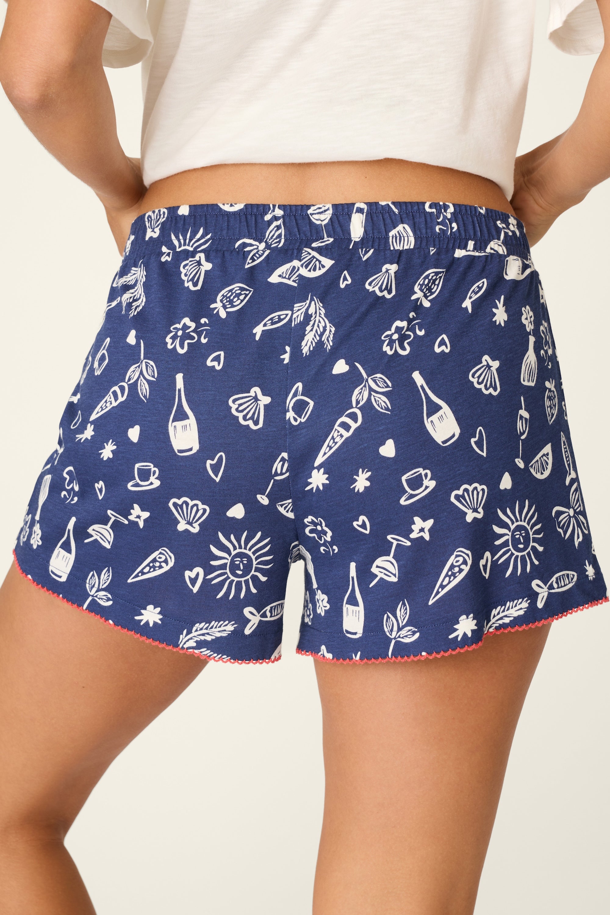 A person wears PJ Salvage Playful Print Boxer Shorts—blue women’s cotton flannel with white wine bottles, carrots, leaves, suns, teacups, and hearts. Features red hem trim and elastic waistband. Waist down shown from the back.