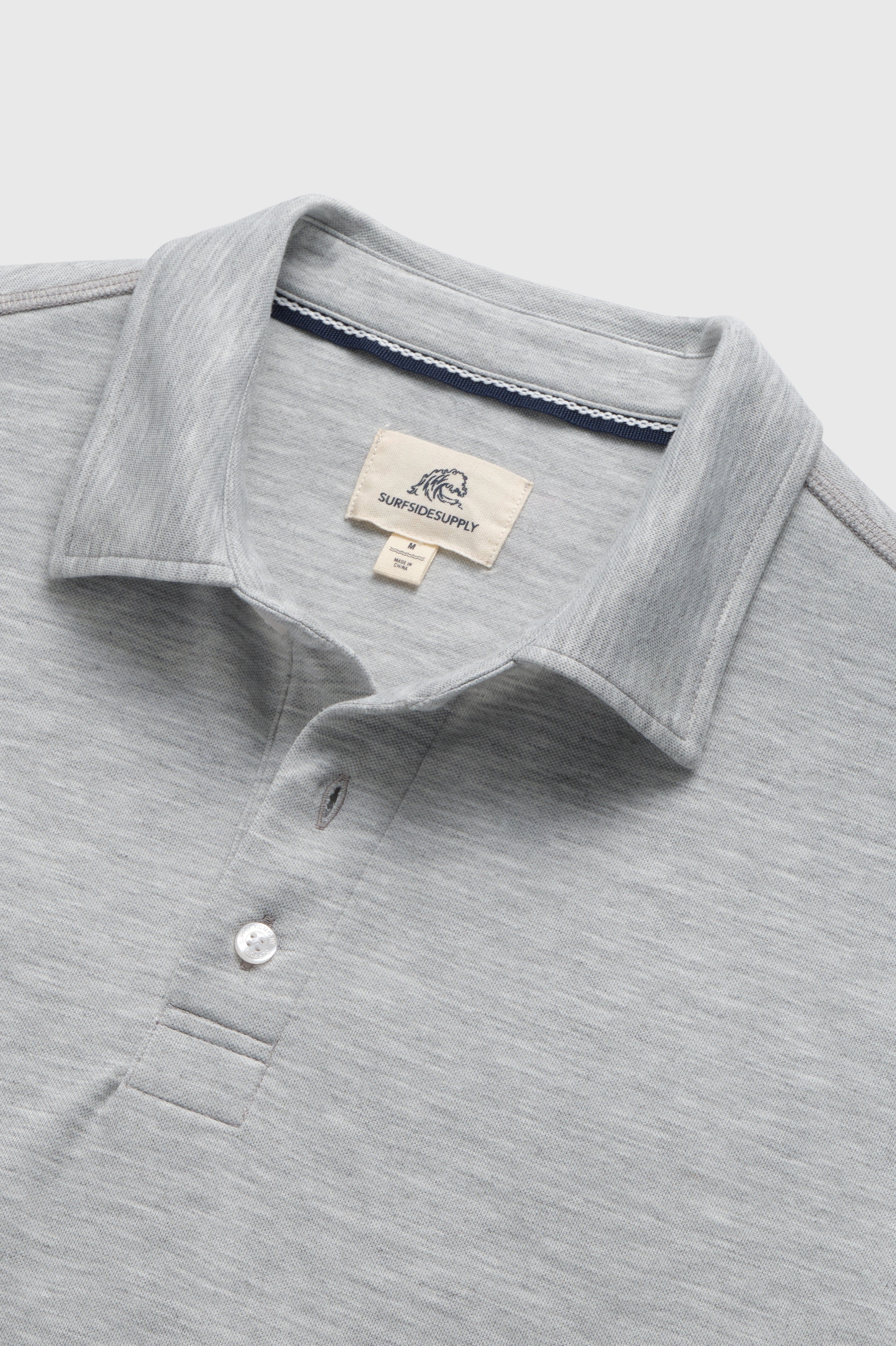 A super soft cotton Ryan Pique Polo from Surfside Supply Co. is shown from the chest up, featuring a light gray color, short sleeves, a collared neckline with two buttons, and a tag inside the collar that reads SUPERICSUPPLY. The polo shirt is laid flat against a plain light background.