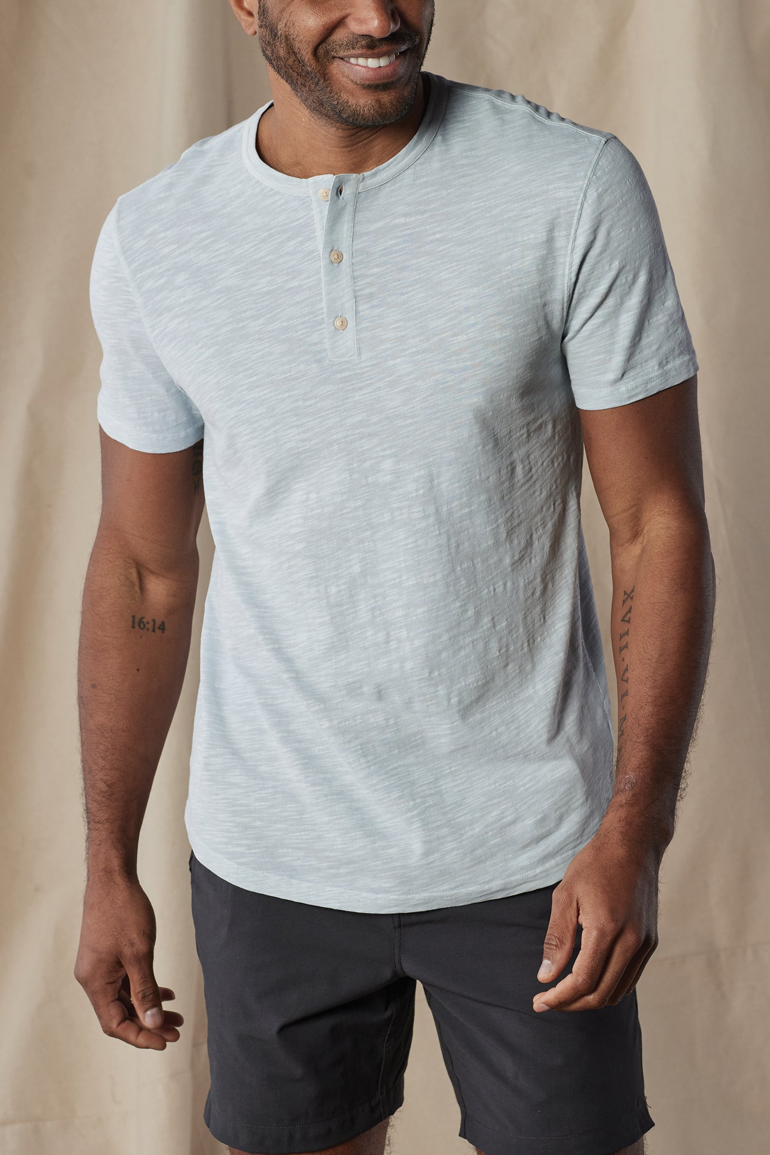 A man in The Normal Brand Legacy Jersey Short Sleeves Henley in light gray and black shorts smiles against a neutral beige fabric backdrop.