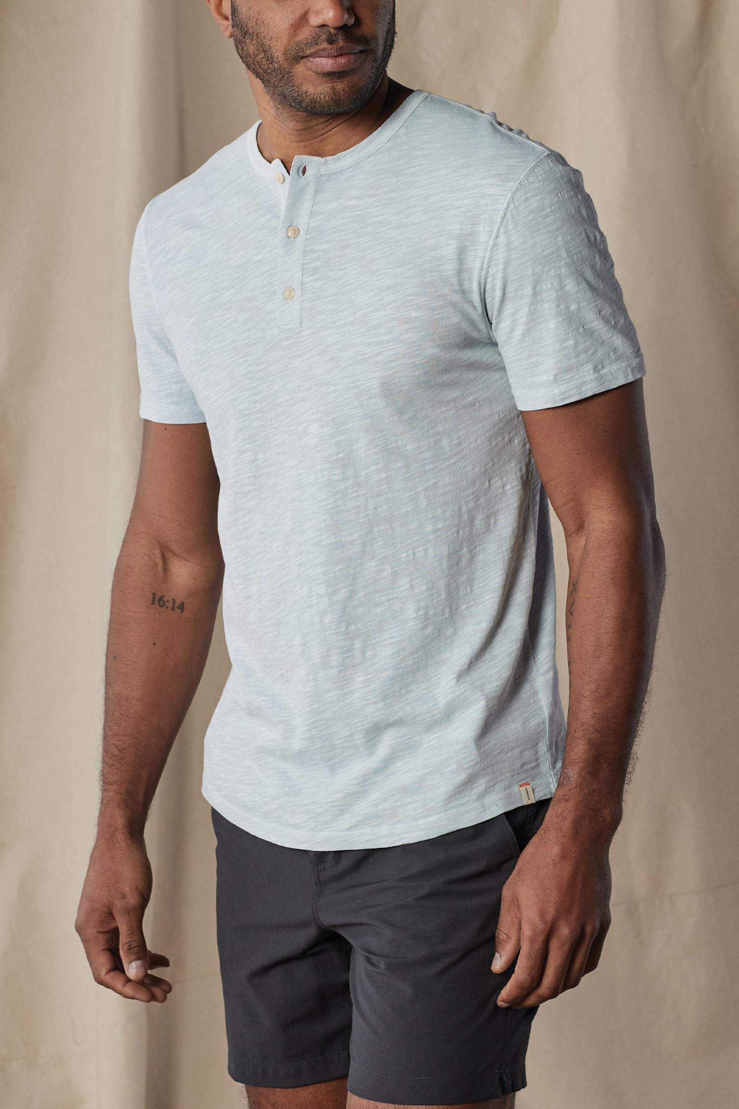 A man stands in front of a beige background, wearing The Normal Brand Legacy Jersey Short Sleeves Henley in light blue and dark shorts, with his arms relaxed by his sides.