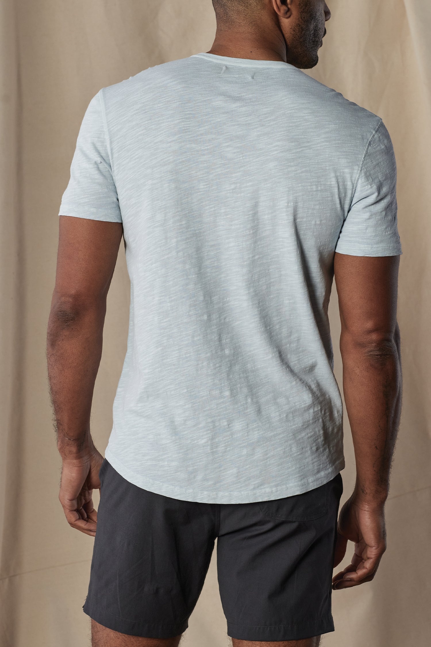 A man faces away from the camera, wearing The Normal Brand's Legacy Jersey Short Sleeves Henley in light blue with dark shorts, set against a beige fabric backdrop.
