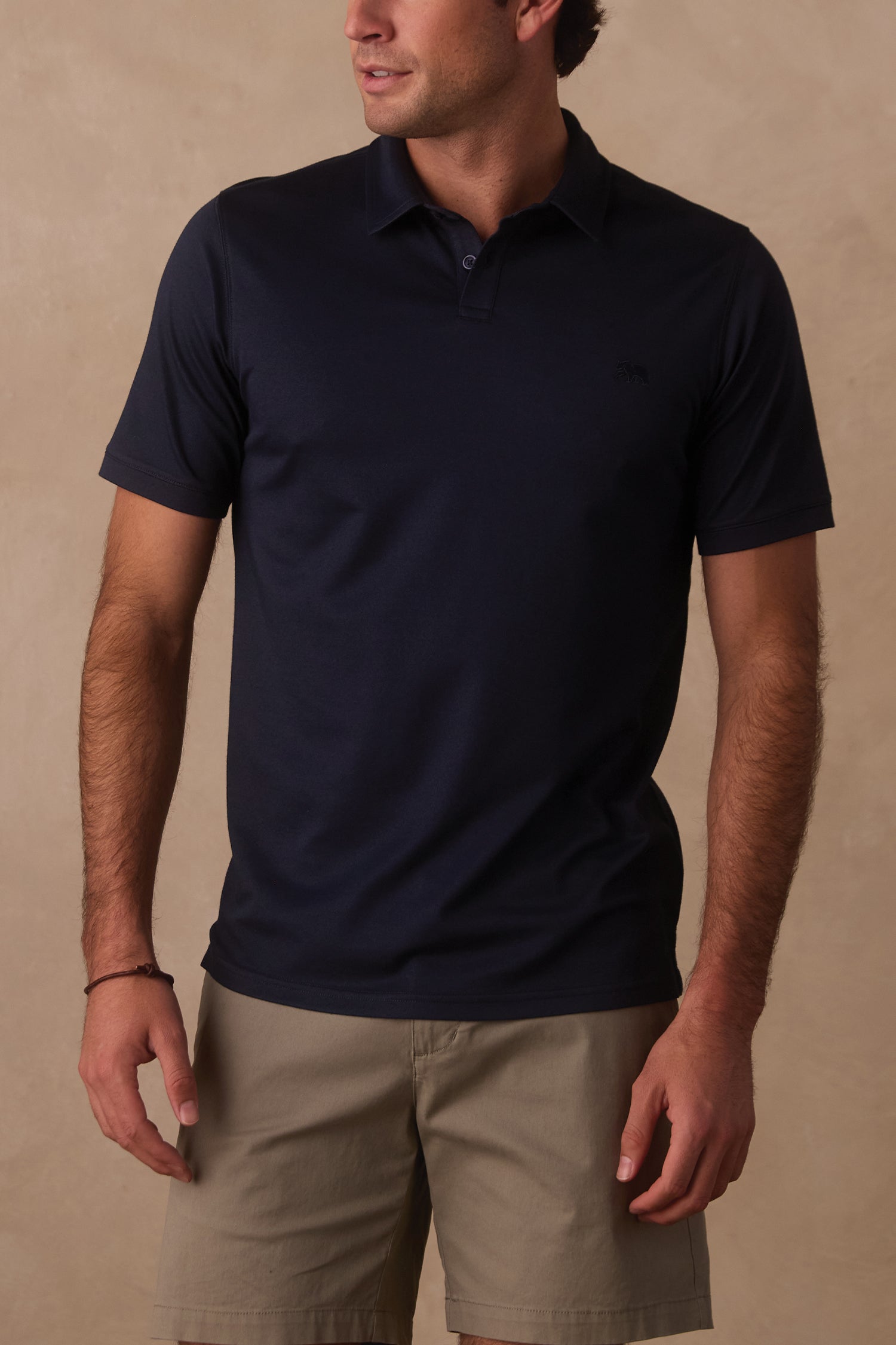 A man models The Normal Brand’s Performance Pique Polo in Navy, standing against a plain light tan background with his hands relaxed at his sides and looking slightly to the side.