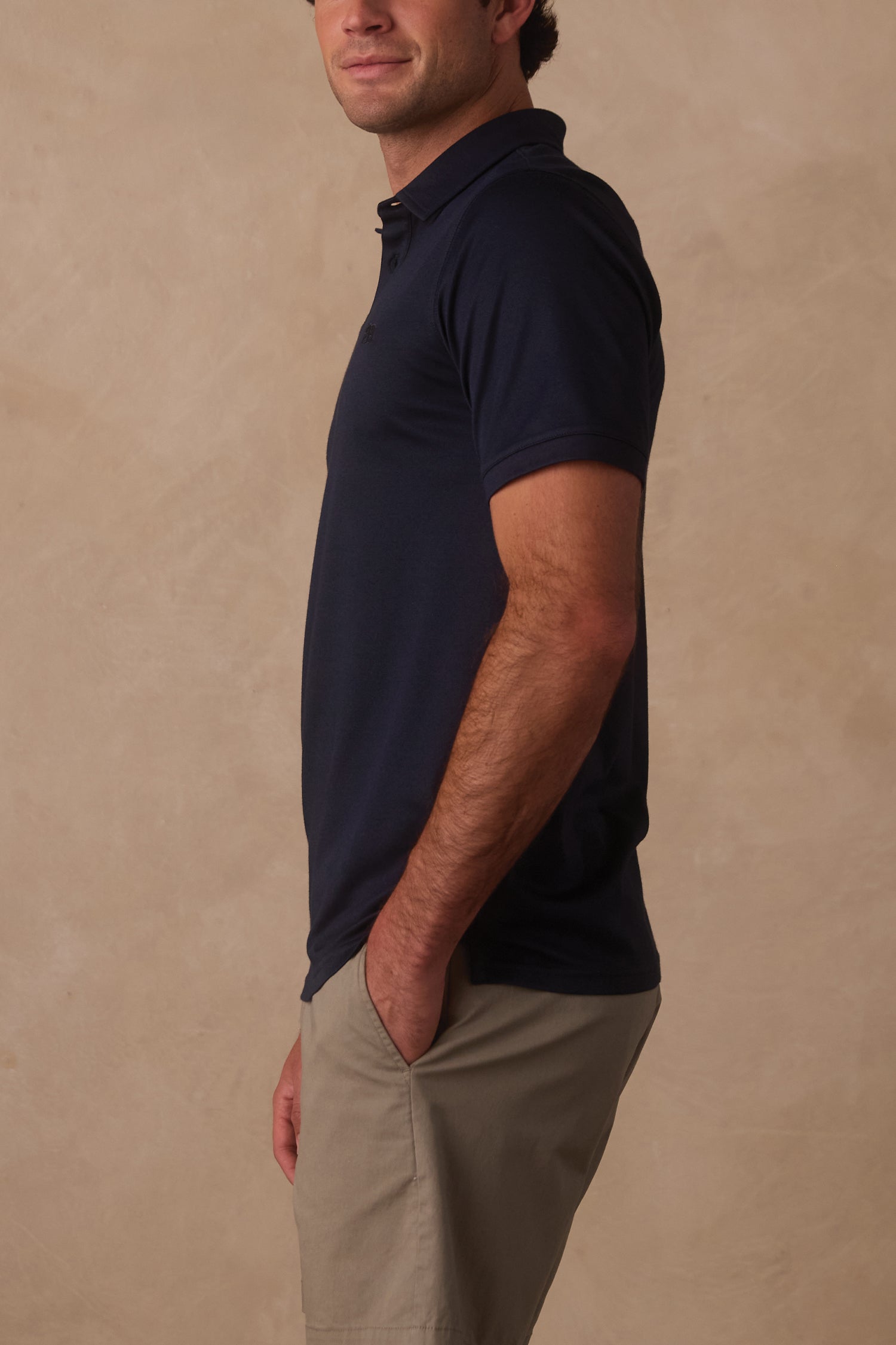 A man stands in profile with one hand in his pocket, wearing The Normal Brand Performance Pique Polo in navy and light khaki shorts against a beige background.