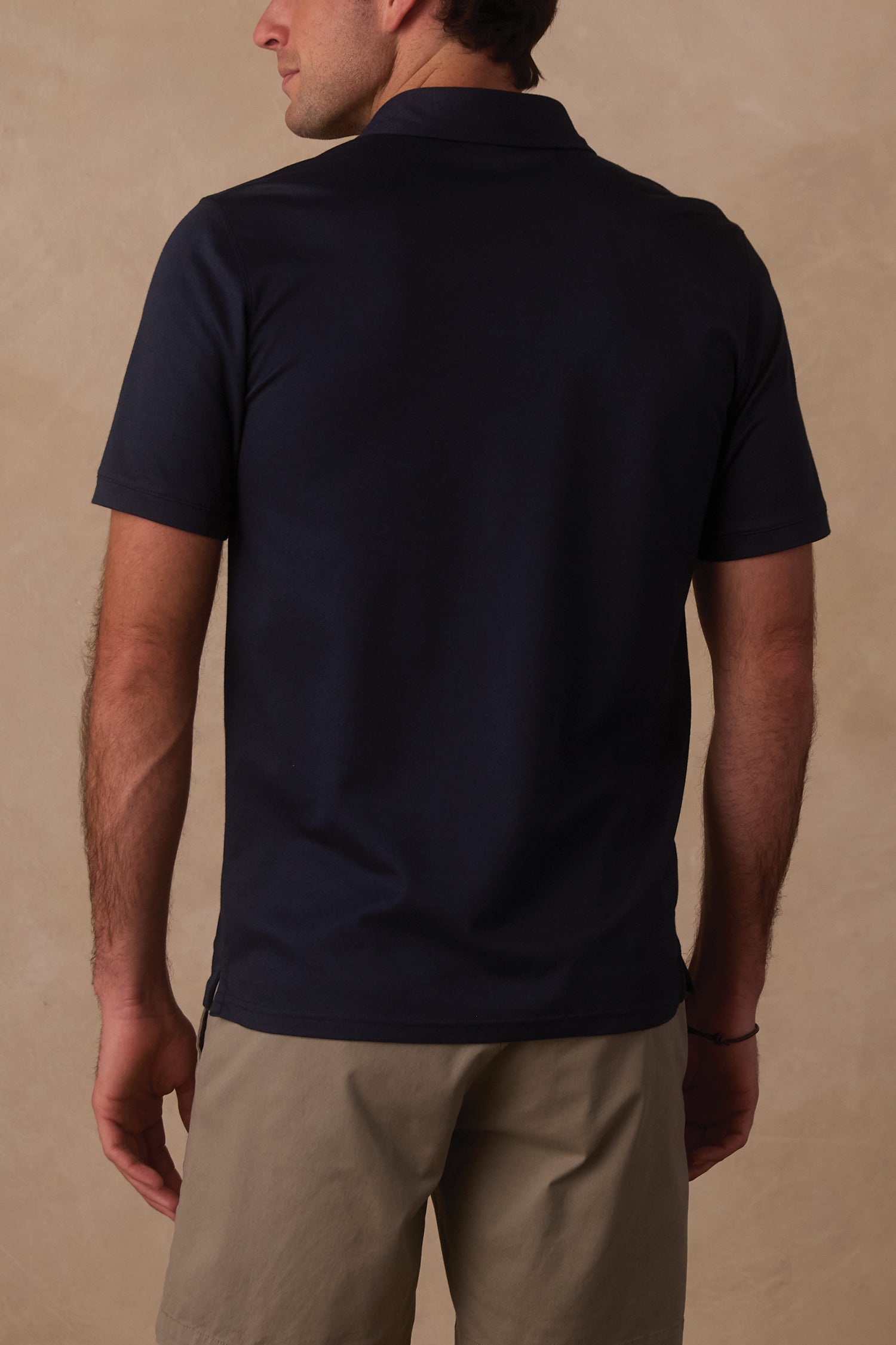 A man with his back to the camera wears The Normal Brand Performance Pique Polo and light khaki shorts, standing against a beige background.
