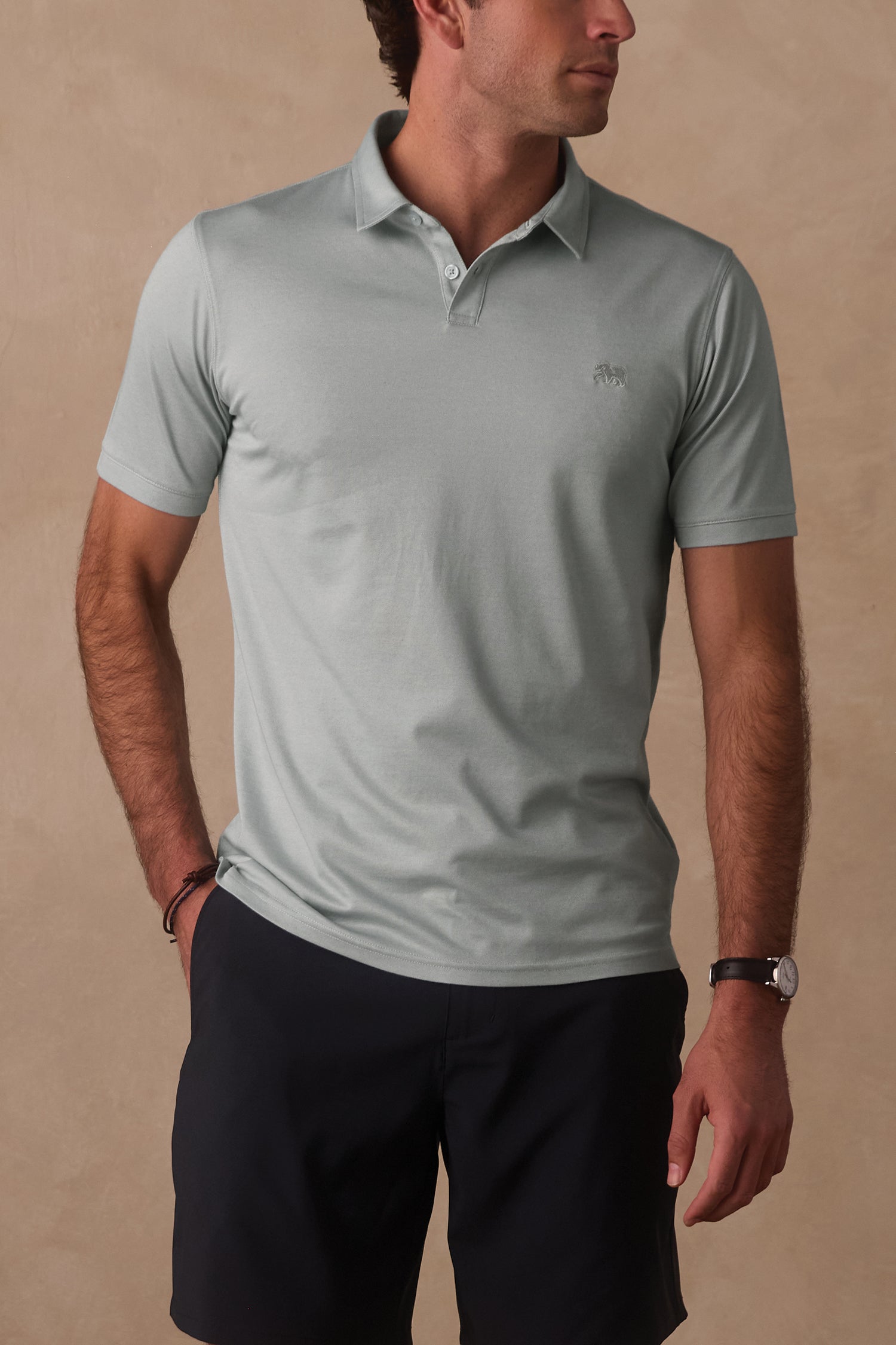 A man wears The Normal Brand’s Performance Pique Polo in light green with black shorts, standing against a beige background and looking to the side with one hand in his pocket.