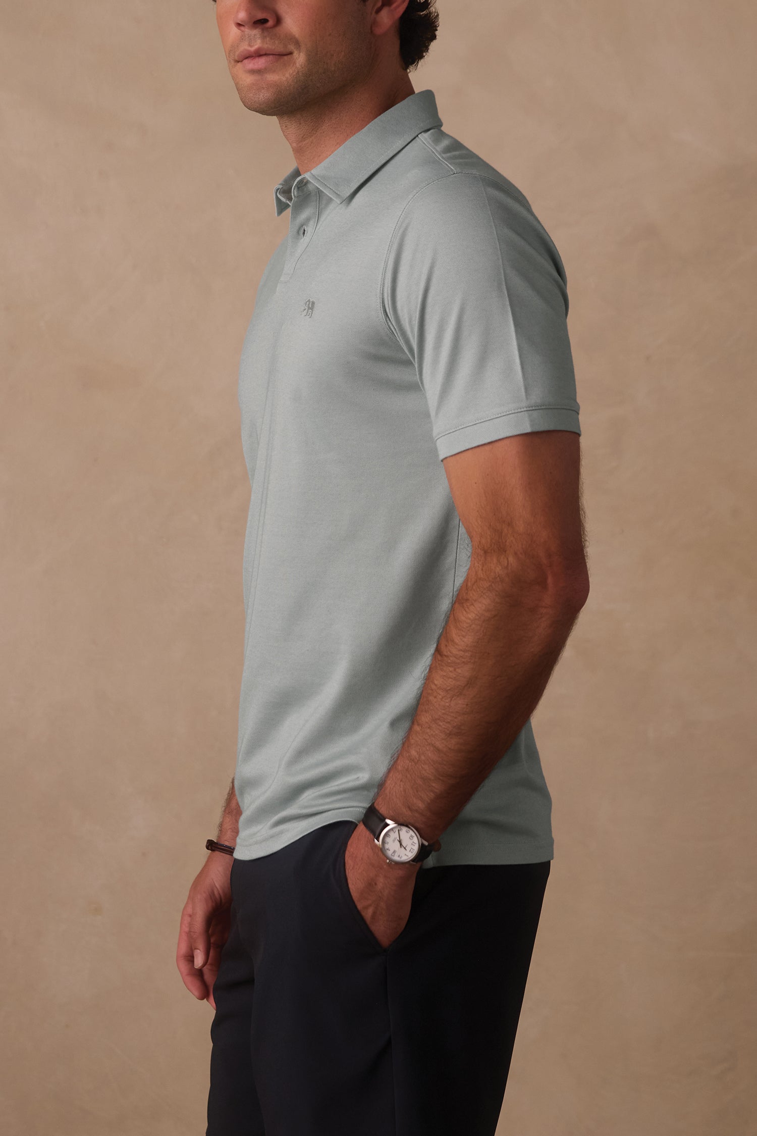A man stands against a beige background with his hands in his pockets, wearing The Normal Brand Performance Pique Polo in light gray and dark pants, facing slightly to the side.