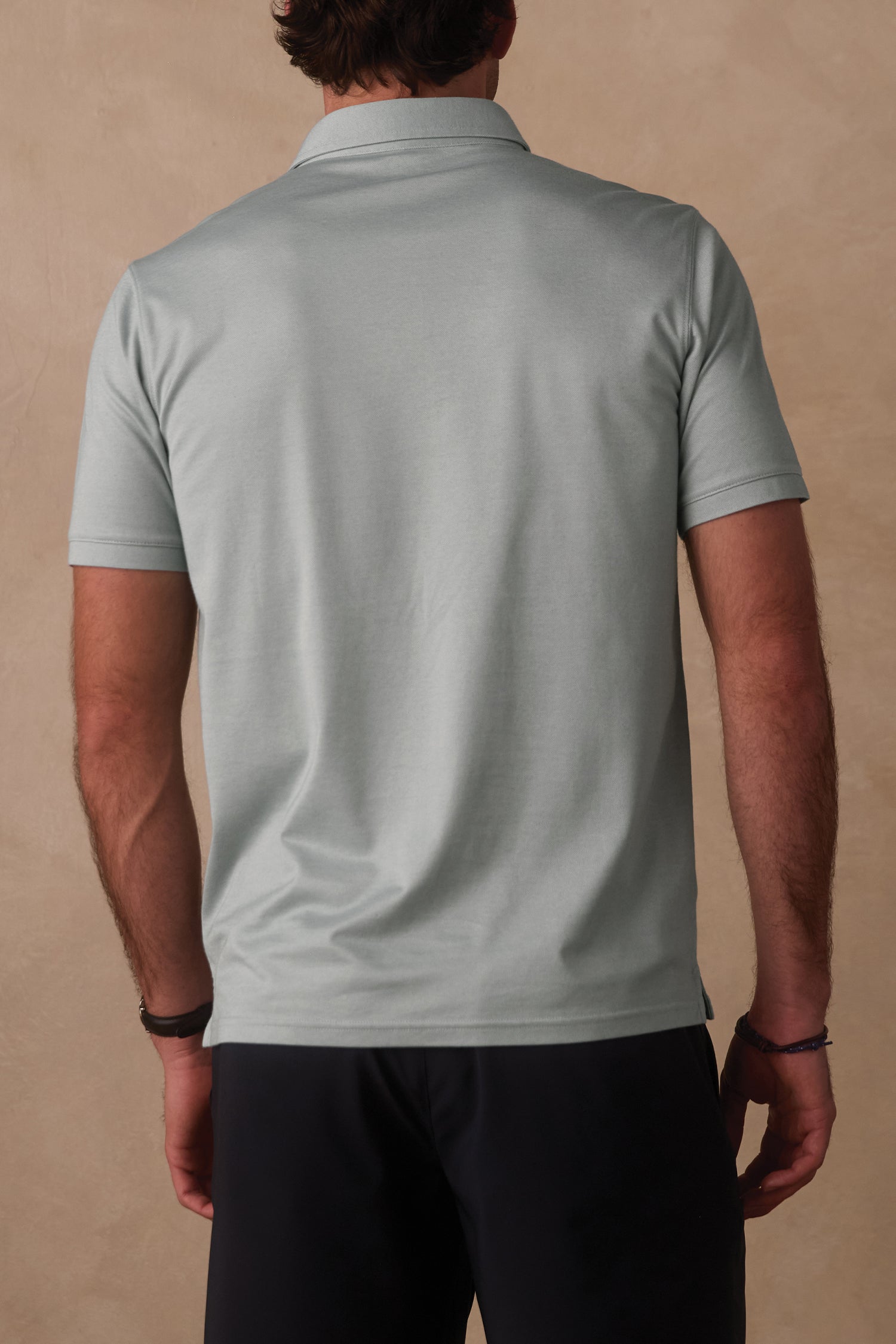 A man is seen from behind wearing The Normal Brand's Performance Pique Polo in light gray with dark pants, standing against a neutral beige background.