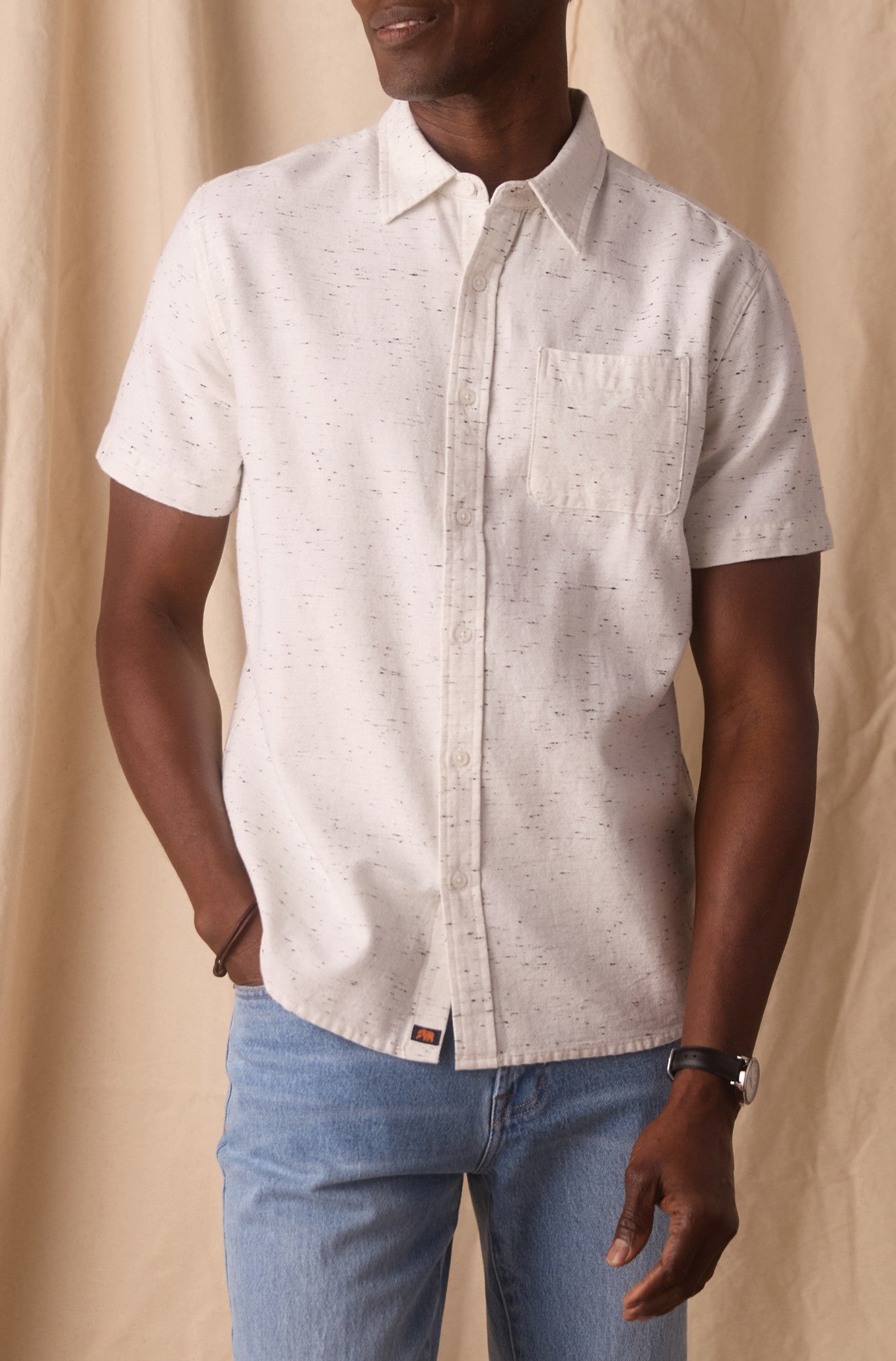 A man wearing The Normal Brand Freshwater Button Up Shirt in WHITE NEP—a short-sleeve, 100% cotton shirt with a chest pocket and durable seams—paired with light blue jeans stands against a beige fabric background.