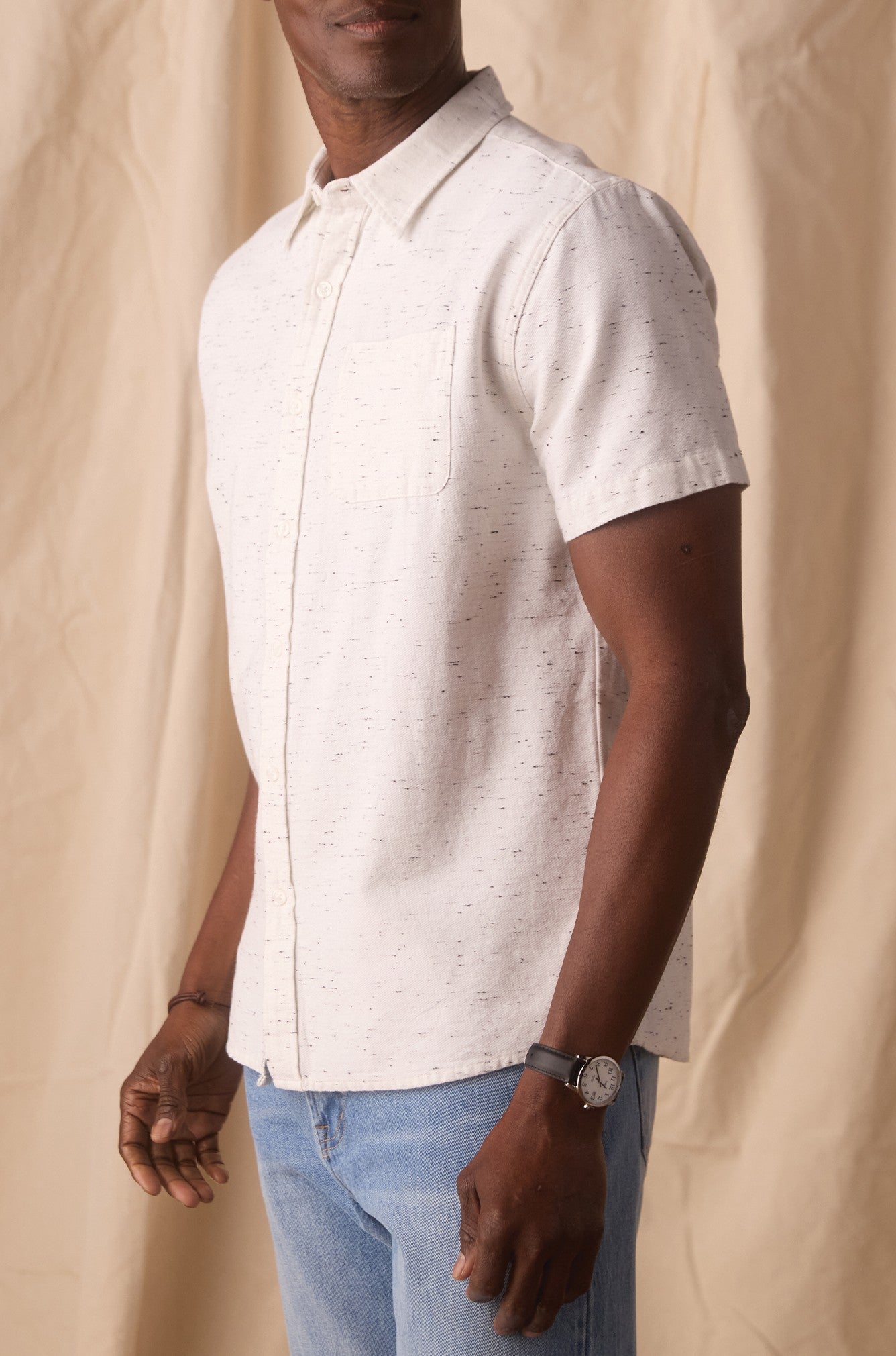 A man stands sideways against a beige fabric background, wearing The Normal Brand's Freshwater Button Up Shirt—a light, short-sleeve button-up made from 100% cotton—paired with blue jeans and a watch on his left wrist.
