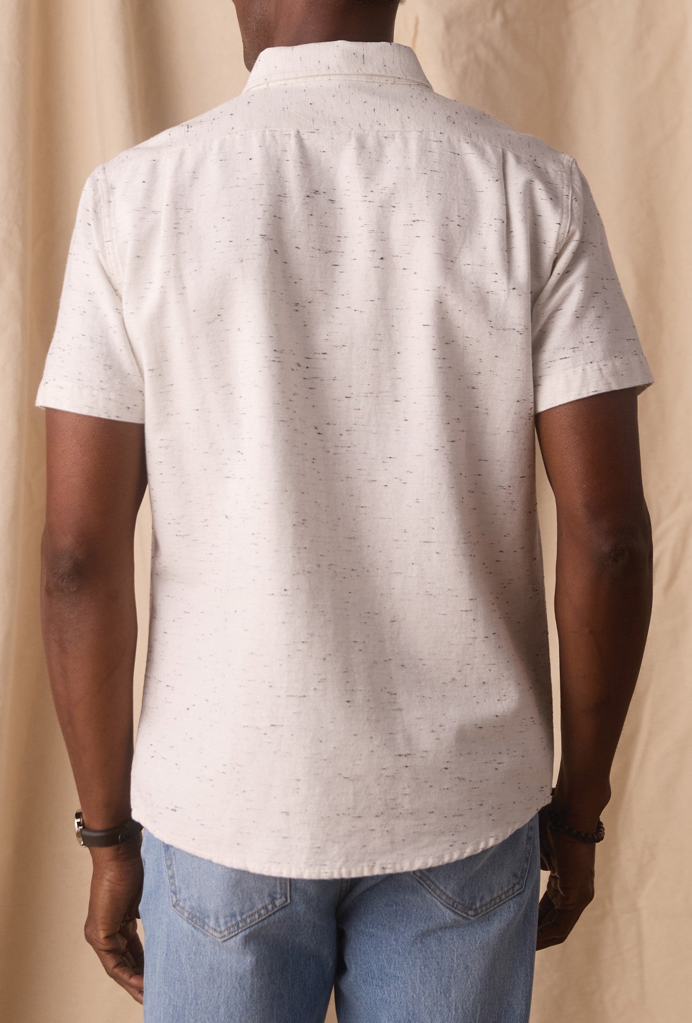 A person is shown from the back, standing against a beige fabric background, wearing The Normal Brand’s Freshwater Button Up Shirt in light gray—a short-sleeve shirt made from 100% cotton with durable seams and a classic button placket—paired with light blue jeans.