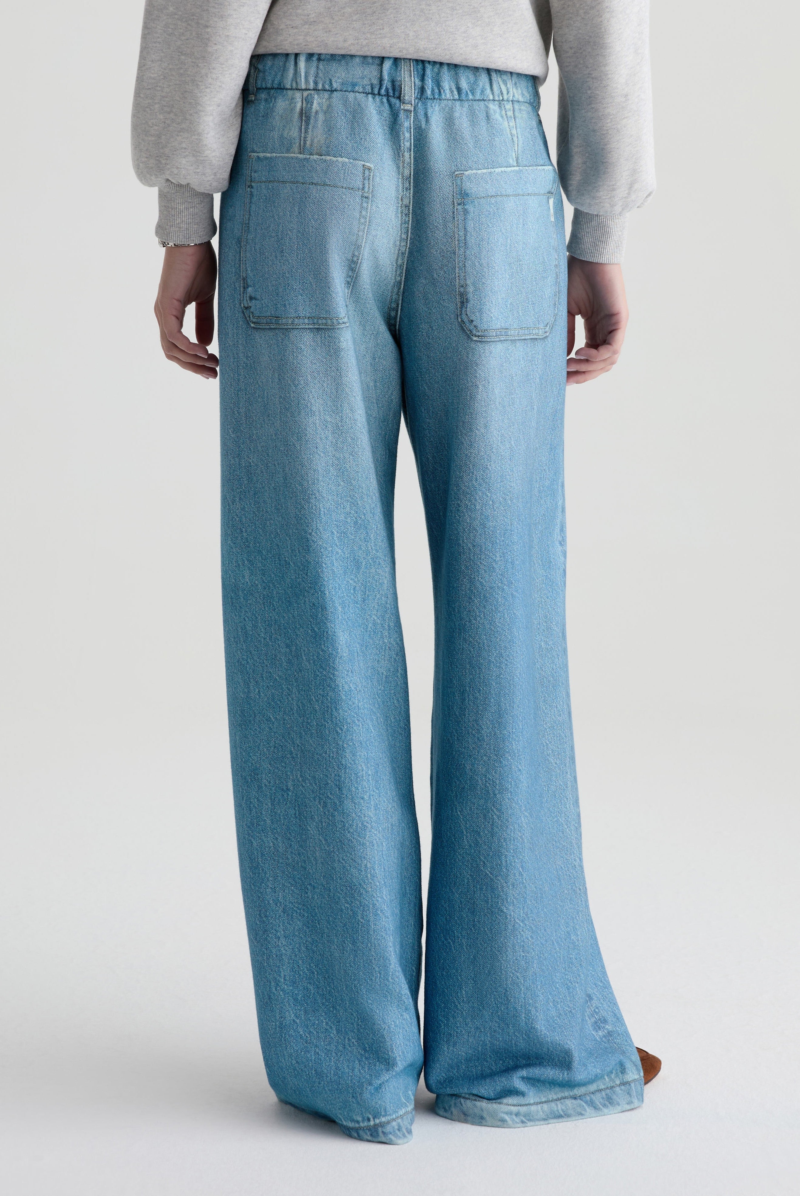A person stands facing away, wearing AG Jeans' Stella Denim Sweatpant—a relaxed fit, high-waisted, wide-leg style with two large back pockets and a light gray sweatshirt. The long jeans nearly touch the floor.