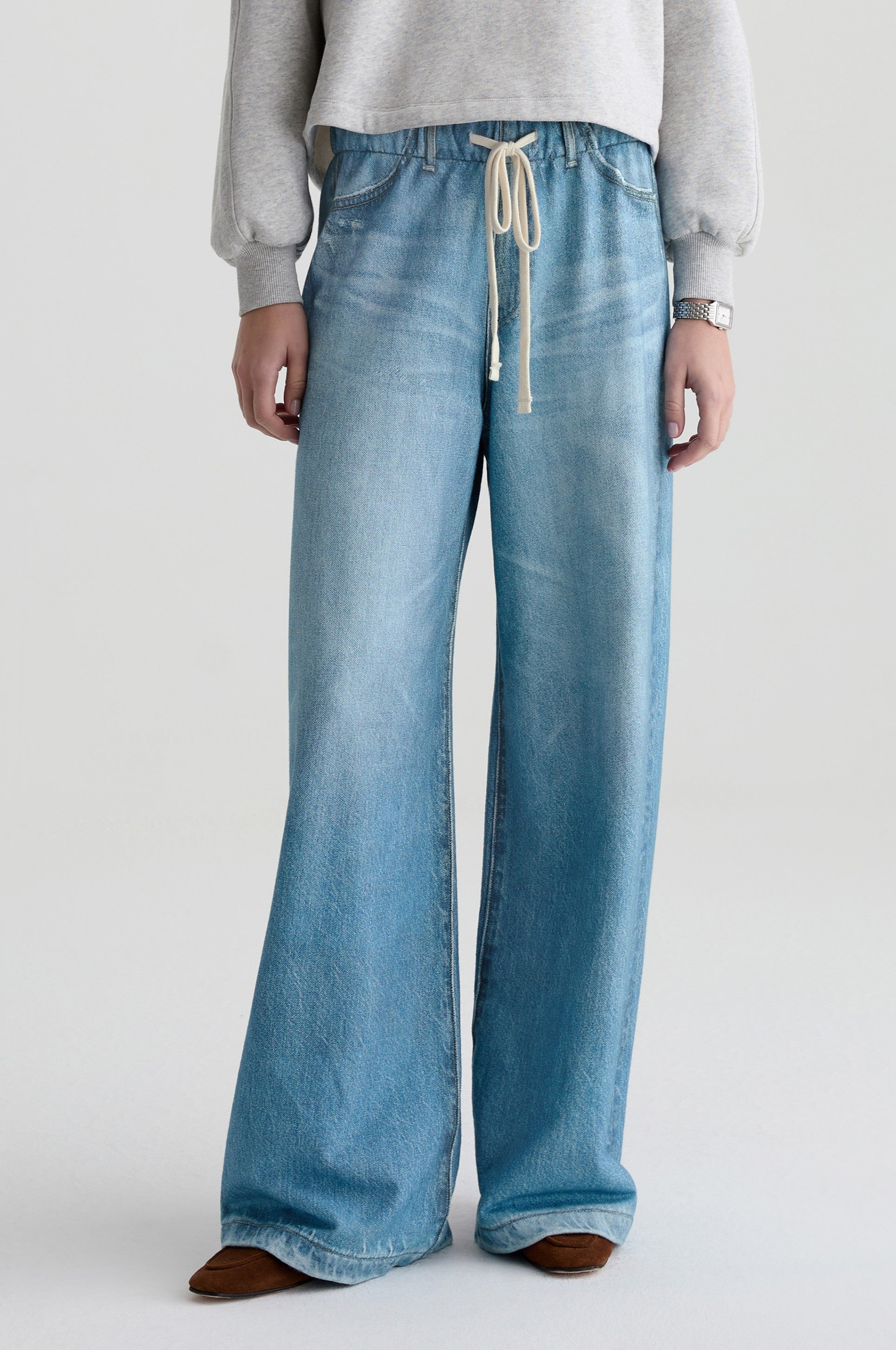 Only the lower body is visible: a person wears the Stella Denim Sweatpant by AG Jeans in denim, paired with a light gray long-sleeve top, watch, and brown pointed shoes.