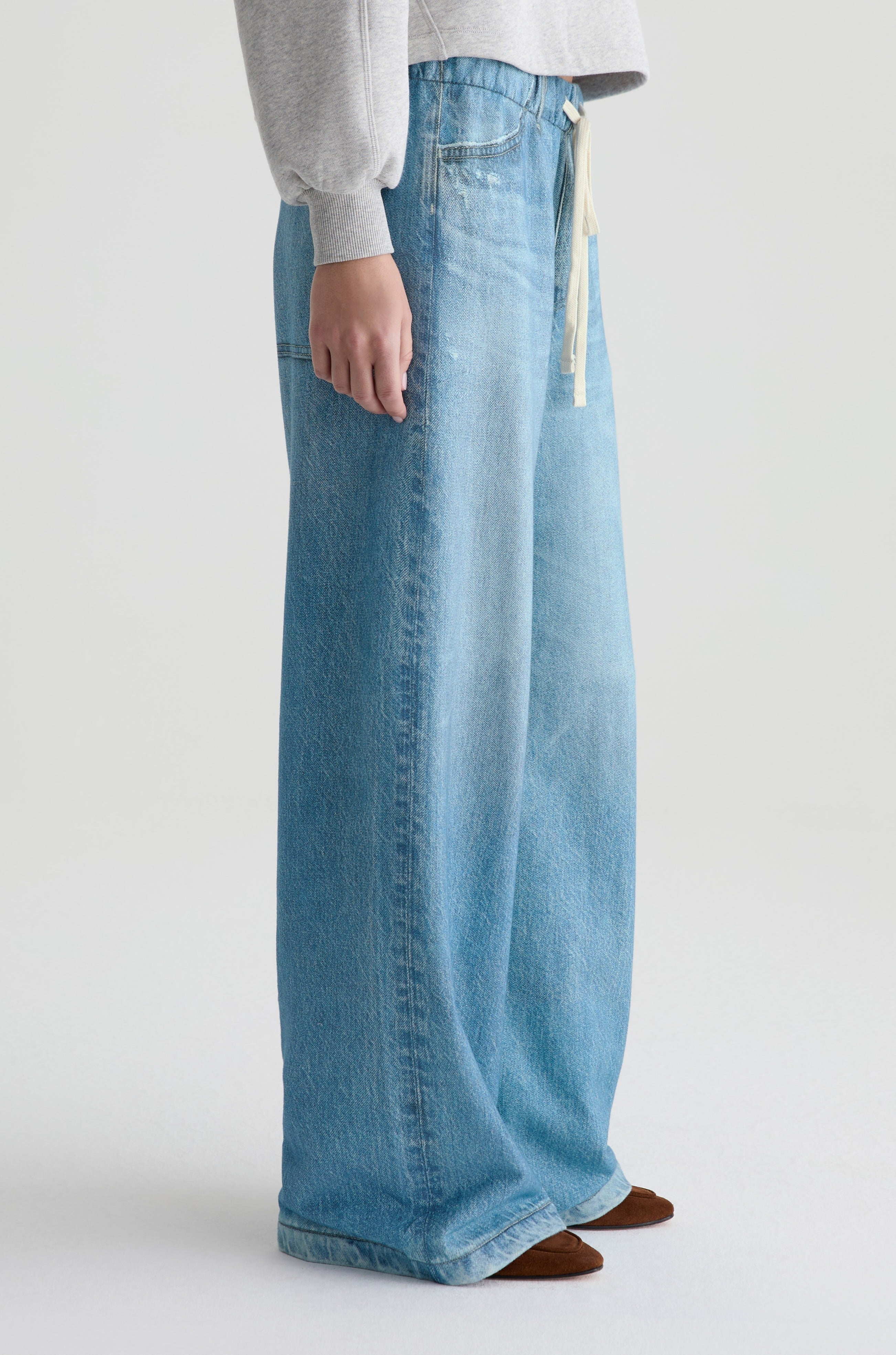 Only the lower half of a person is visible, wearing AG Jeans' Stella Denim Sweatpant in light blue with a relaxed fit and drawstring waistband, paired with a grey sweatshirt, standing against a white background.