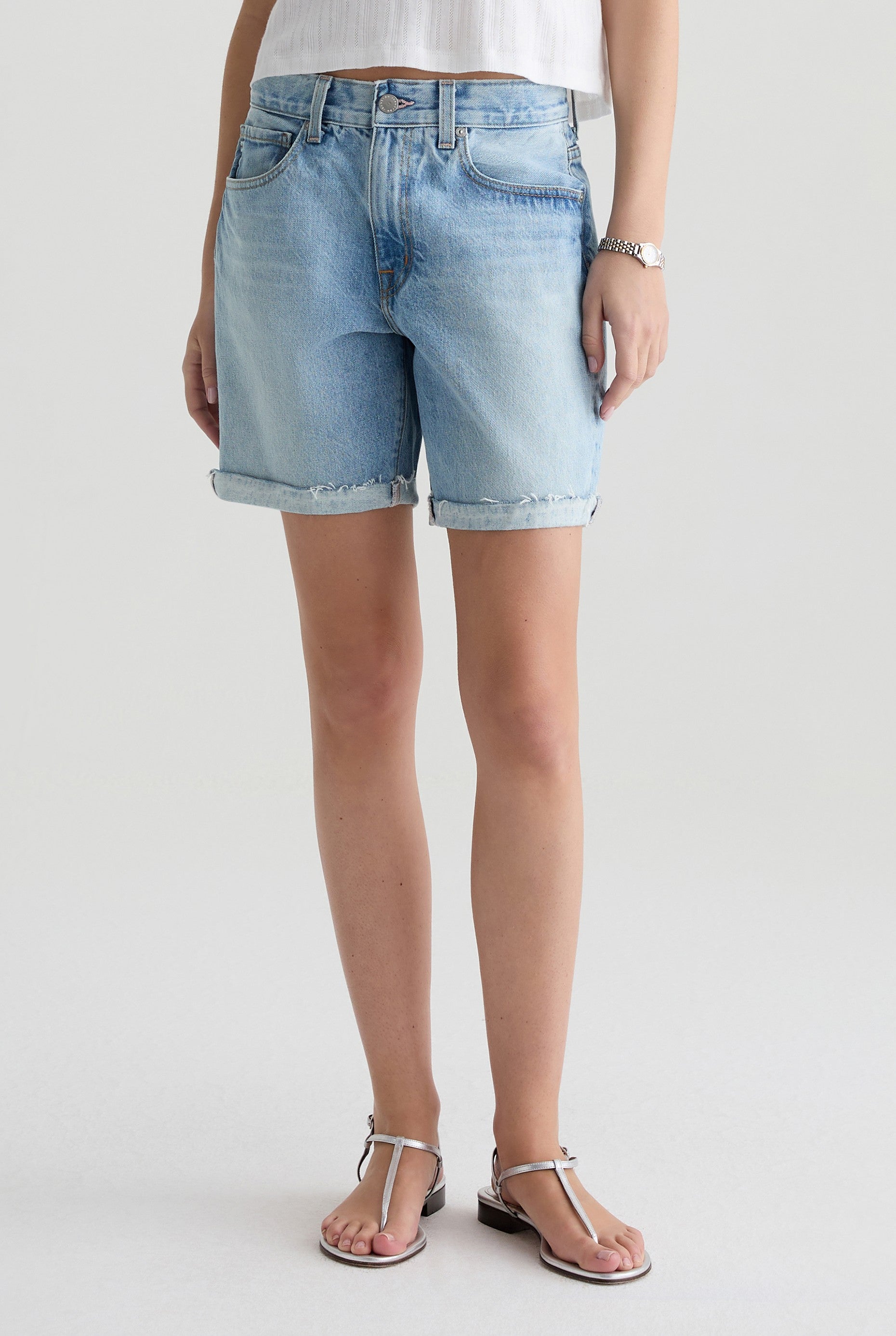 A person wearing AG Jeans' Ex-boyfriend Short in light blue rigid 100% cotton denim, paired with a white sleeveless top, silver bracelet, and silver sandals stands against a plain light background.