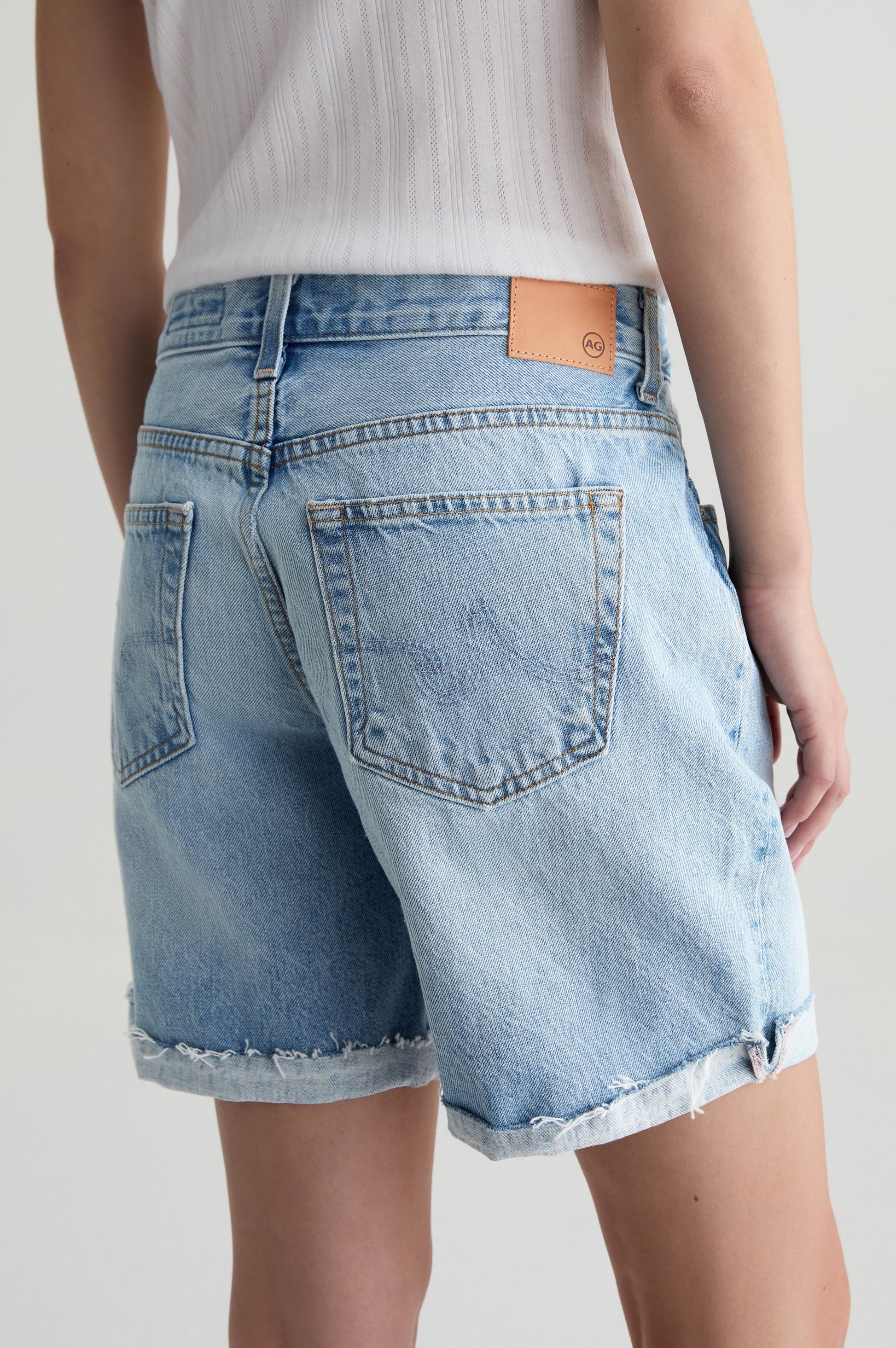 A person wearing AG Jeans’ Ex-boyfriend Short: light blue, high-waisted, 100% cotton rigid denim shorts with frayed hems, two large back pockets, and a tan leather patch, paired with a white ribbed sleeveless top. Shown from the back.
