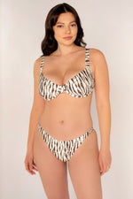 A woman with long brown hair models the Amanda Batula Swim Snake Underwire Top, a cream and brown patterned bikini with a tie back, standing against a plain light background and smiling slightly at the camera.