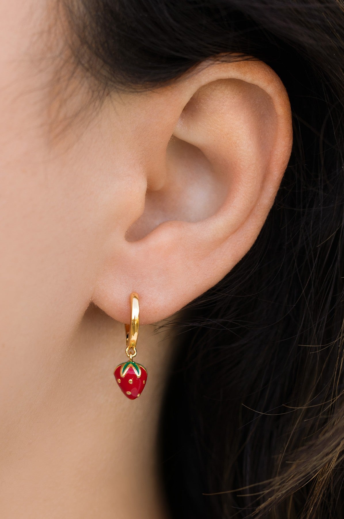 A close-up of a person’s ear wearing Girls Crew Enamel Strawberry Mini Hoops in red, with the charming strawberry detail peeking out from beneath dark hair.