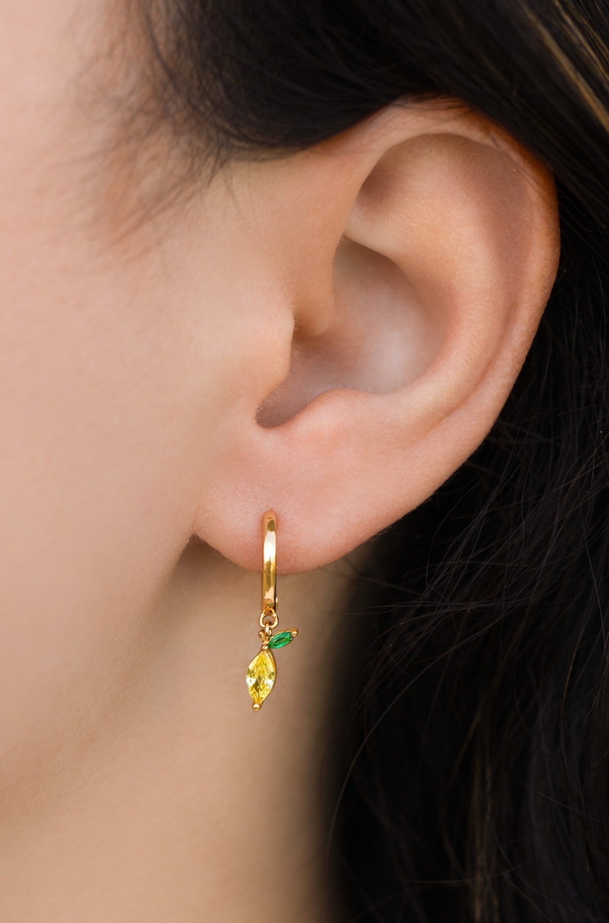 A close-up of an ear wearing Girls Crew Stone Lemon Mini Hoops: yellow drop earrings featuring a small gold-tone huggie with a leaf-shaped gemstone and a green accent stone.