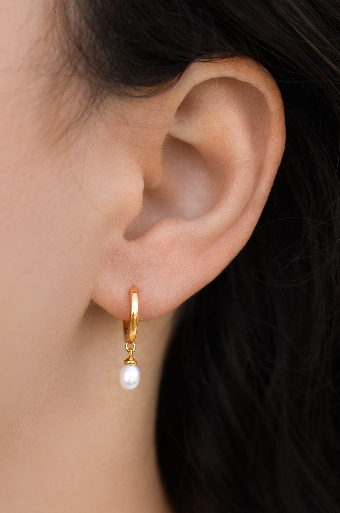Close-up of a person’s ear wearing South Moon Under’s Pearl Mini Hoops—delicate gold earrings each with a single white pearl drop. The blurred background draws attention to these elegant Pearl Mini Hoops.