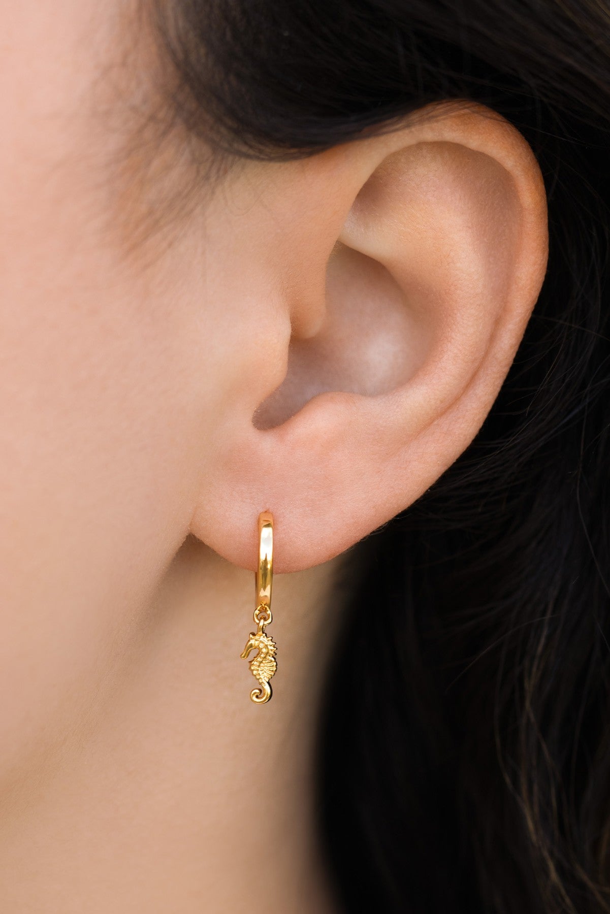 A close-up shows a light-skinned ear wearing Girls Crew’s gold Simple Seahorse Hoops—small huggie earrings with a dangling seahorse charm—paired with dark hair for a subtle, coastal-inspired style.