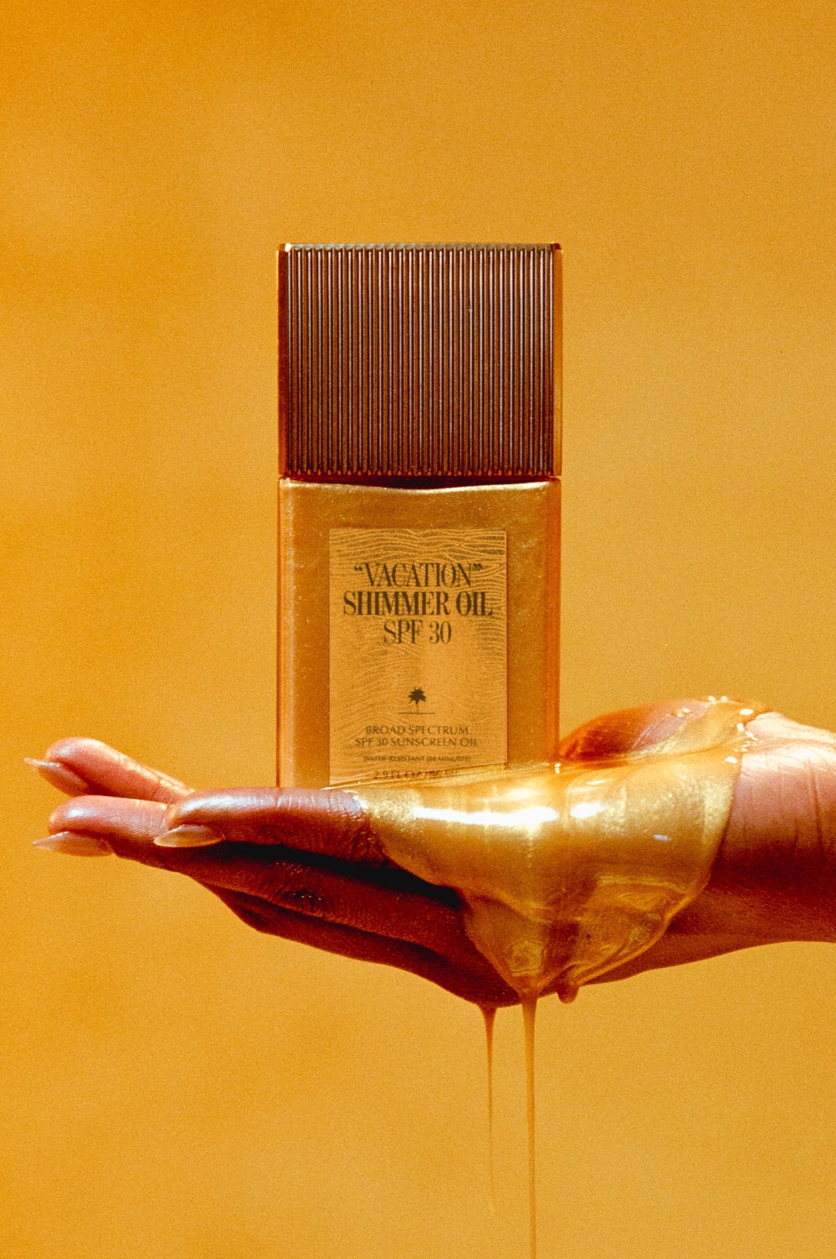 A hand, coated in golden shimmer oil, holds a bottle of Vacation Shimmer Oil SPF 30 by Vacation against a warm, golden backdrop.