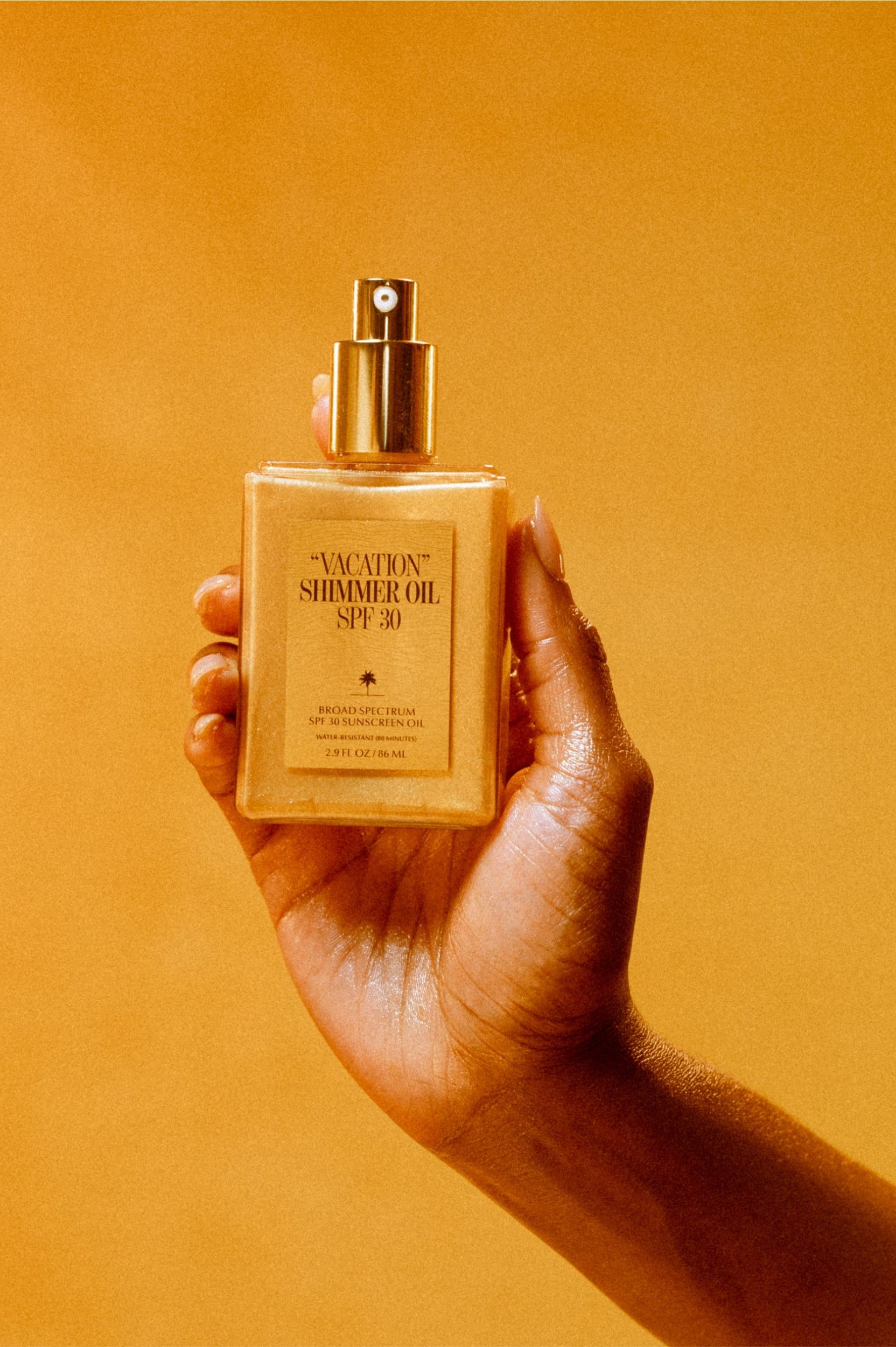 A hand holds a rectangular Vacation Shimmer Oil SPF 30 bottle with a gold pump, featuring broad spectrum protection and coconut oil, set against a golden yellow background.