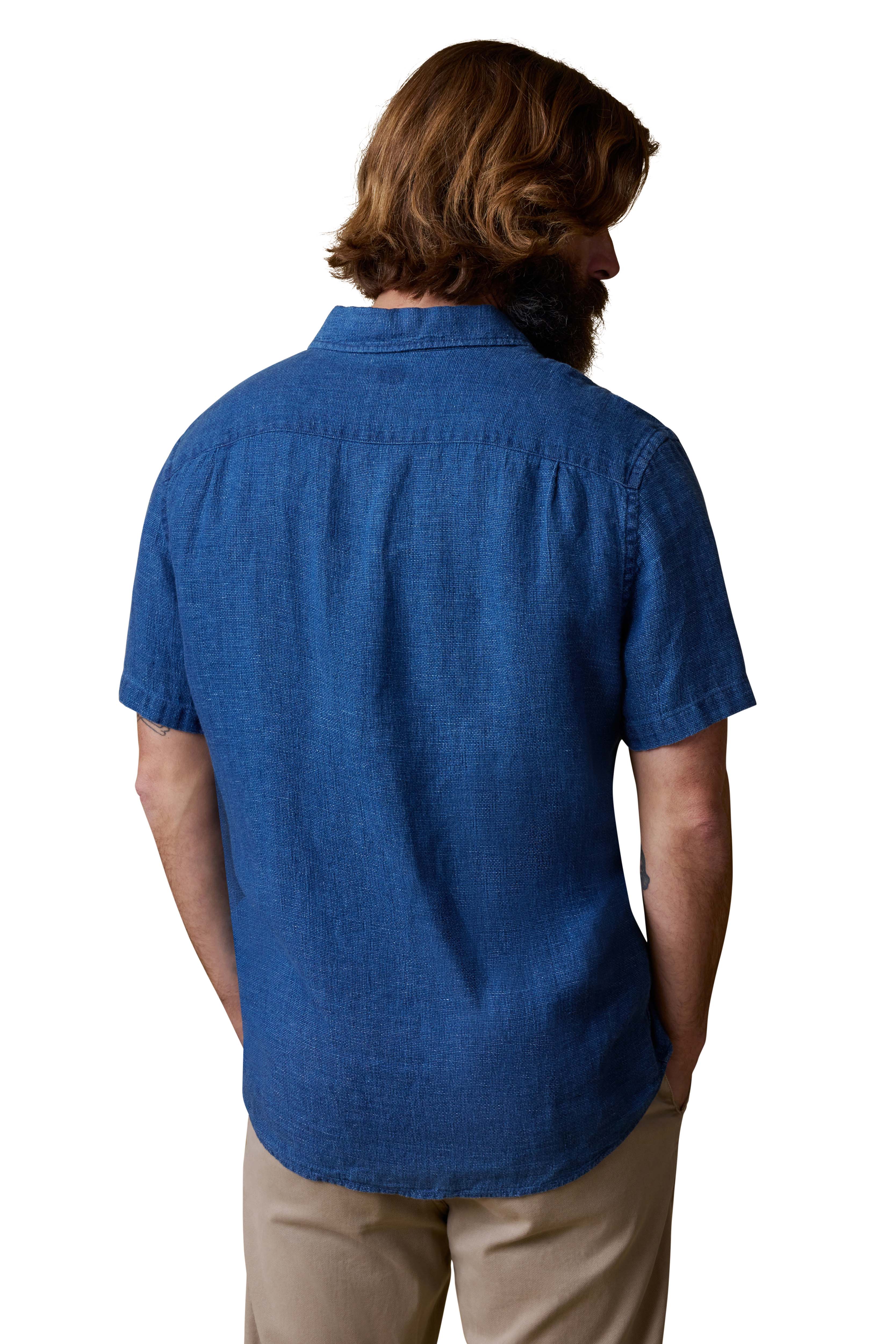 A man with medium-length brown hair and a beard is seen from behind, wearing the Faherty Short Sleeve Palma Linen Shirt in blue and beige pants, set against a white background.