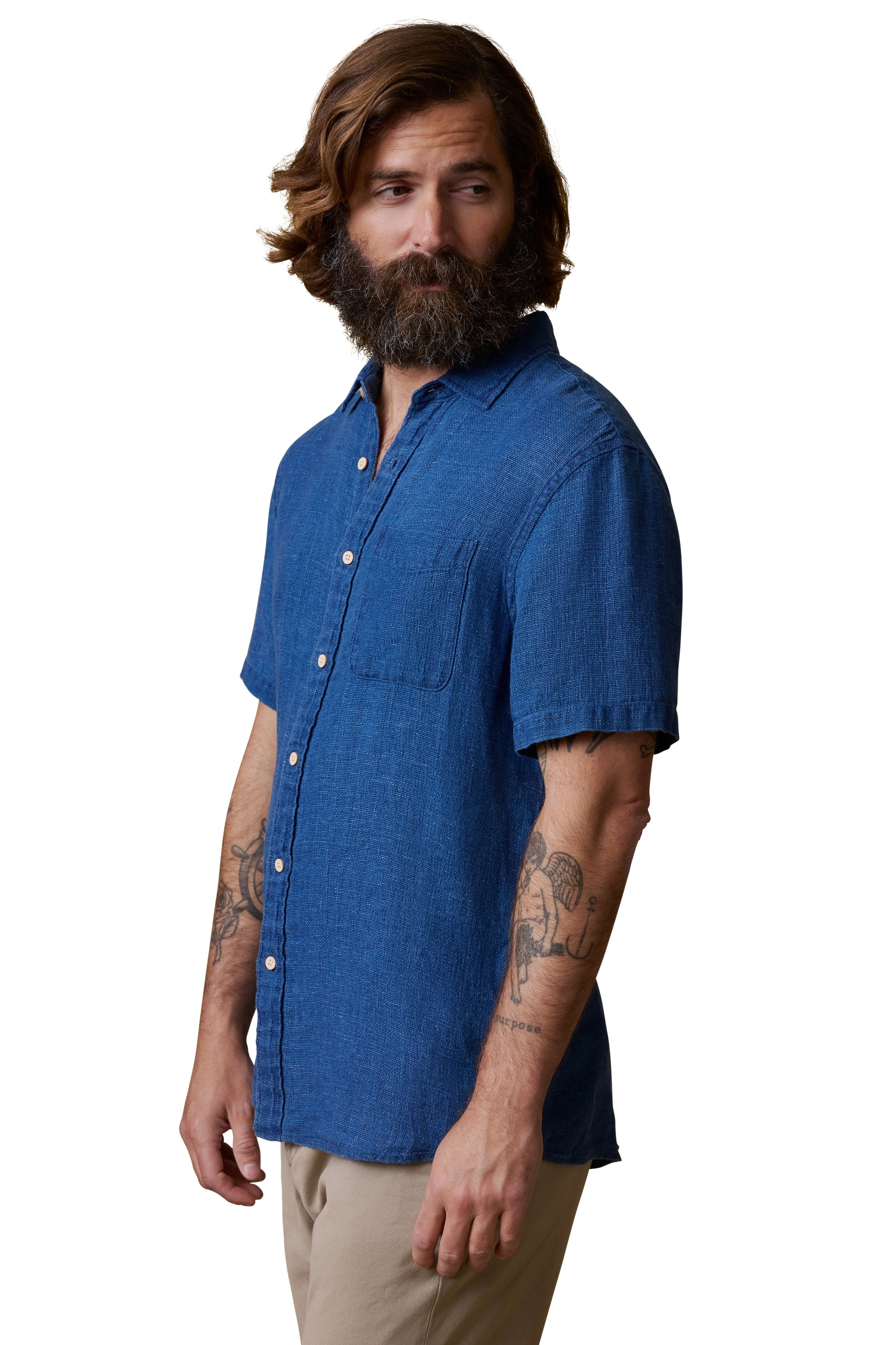 A bearded, tattooed man stands looking left against a white background, wearing the Faherty Short Sleeve Palma Linen Shirt in blue with a button front and straight fit, paired with beige pants.