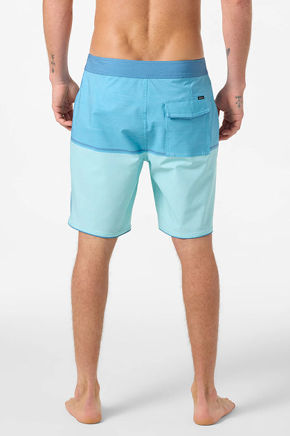 A man stands barefoot, facing away, wearing O'Neill Hyperfreak Mysto Block 19 In Boardshorts in light blue with a dark blue waistband and back pocket. His arms are relaxed by his sides, showing small forearm tattoos. Made from recycled polyester.