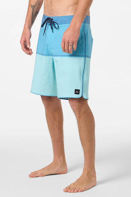 A man stands barefoot in O'Neill Hyperfreak Mysto Block 19 In Boardshorts, made from recycled polyester, featuring a two-tone dark and light blue design, set against a plain white background.